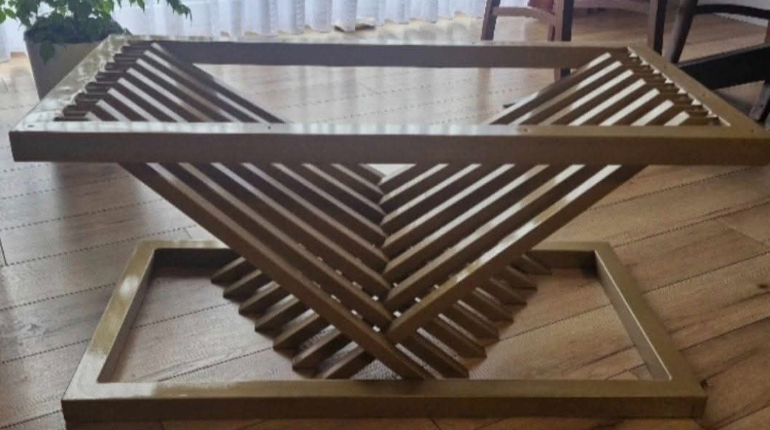 Stolik kawowy ze złotego metalu o geometrycznej konstrukcji, z kwadratowym blatem i podstawą połączoną ukośnymi listwami, na drewnianej podłodze.