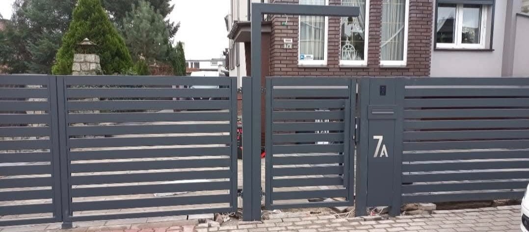 Nowoczesna, pozioma brama wjazdowa w kolorze grafitowym z numerem domu '7A', zintegrowana ze skrzynką na listy i oświetleniem, na tle domu z cegły.