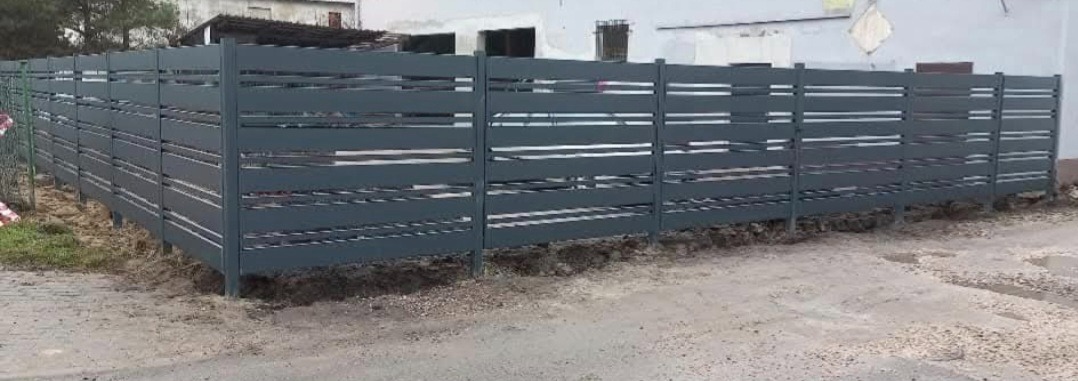 Nowoczesne ogrodzenie metalowe w kolorze grafitowym z poziomymi, wąskimi panelami, osadzone na ziemi przy budynku. Widok z zewnątrz posesji.