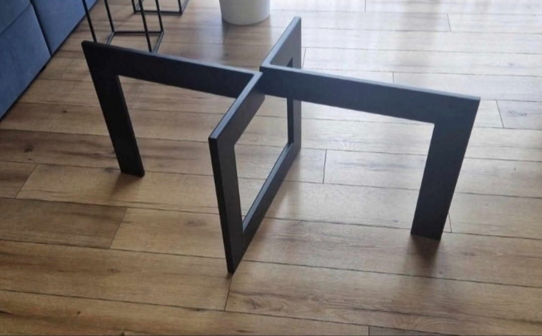 Czarna, geometryczna podstawa stolika kawowego z metalowych profili na drewnianej podłodze. Minimalistyczny design.