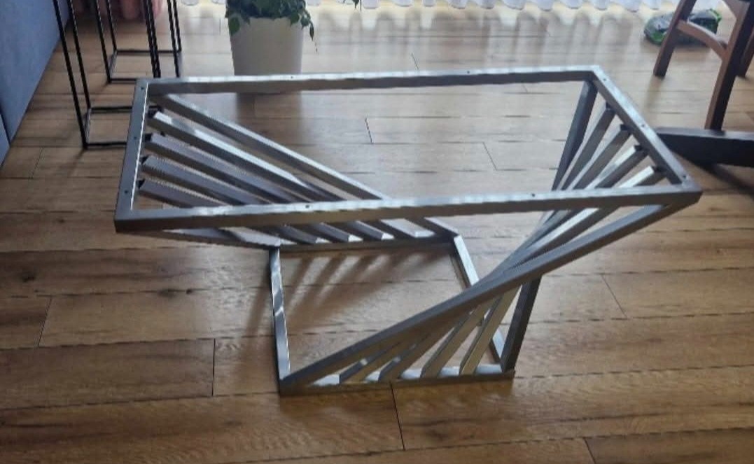 Metalowy stelaż stolika kawowego o geometrycznej konstrukcji, spawany z profili. Widoczne spawy, surowy wygląd metalu, drewniana podłoga w tle.