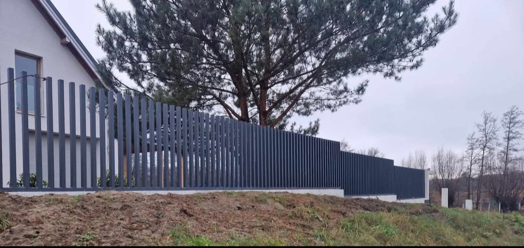Nowoczesne ogrodzenie metalowe z pionowych, wąskich profili w kolorze grafitowym, na tle domu i drzewa iglastego. Widok z poziomu gruntu, pochmurne niebo.
