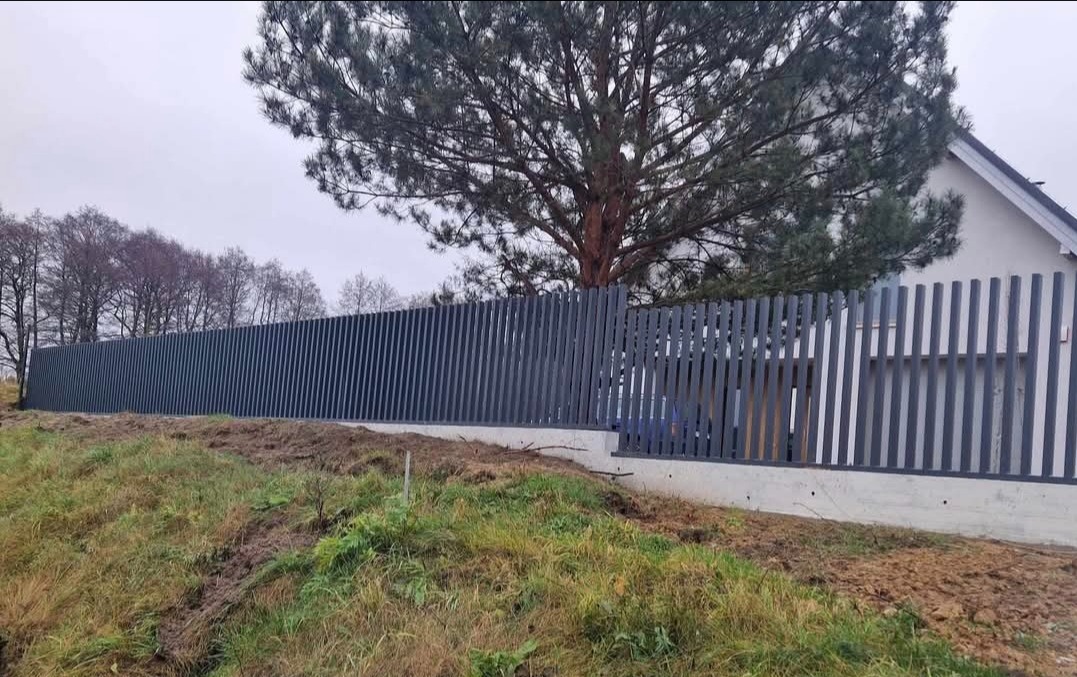 Długie, szare ogrodzenie panelowe z pionowych listew, zamontowane na betonowej podmurówce, oddziela posesję od terenu zielonego z drzewami w tle.