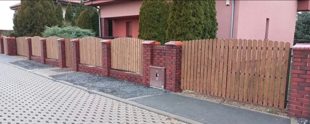 Nowoczesne ogrodzenie drewniane z ceglanymi słupkami i podmurówką, oddzielające posesję od chodnika z kostki. Widoczne elementy architektury domu w tle.