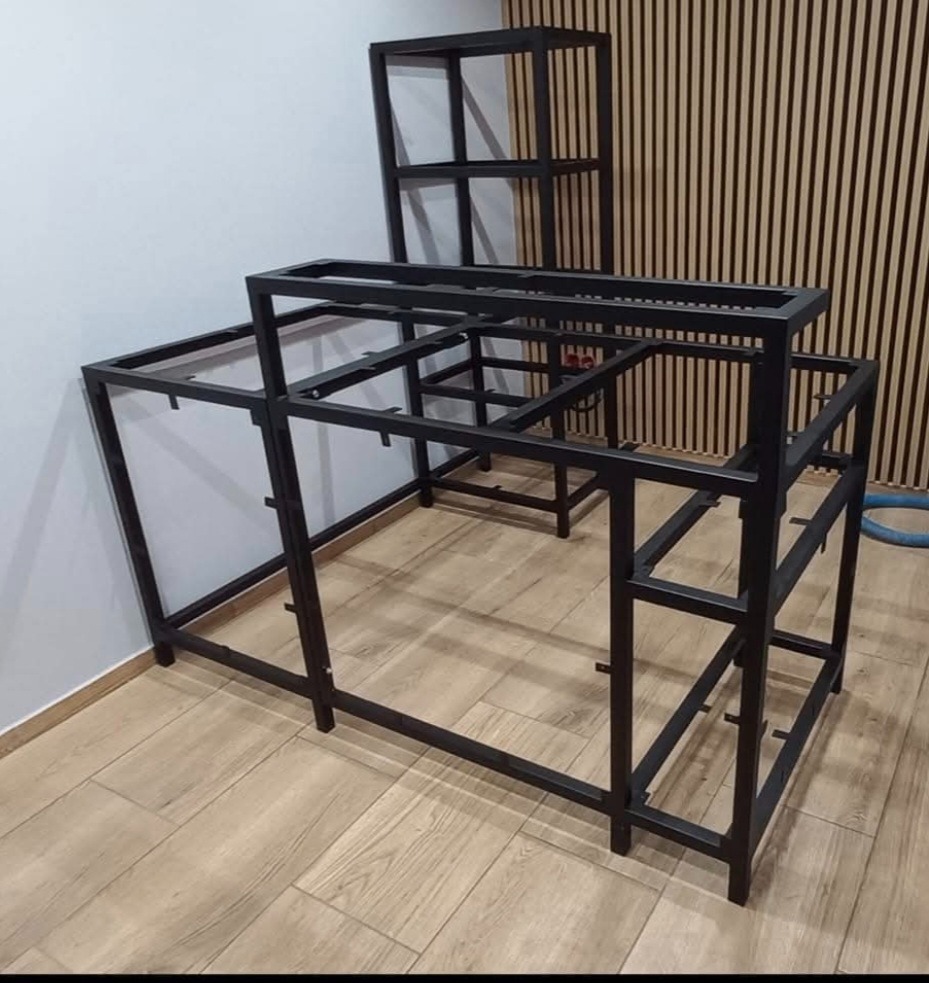 Czarna, metalowa konstrukcja meblowa w surowym stanie, prawdopodobnie do zabudowy baru lub lady recepcyjnej, z dodatkowym regałem. Widoczne spawy i surowe wykończenie.