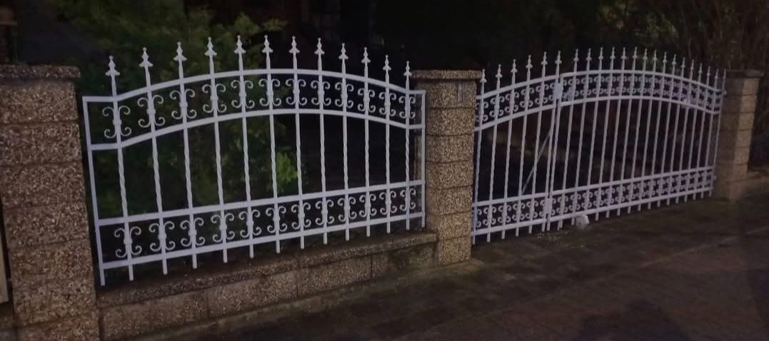 Białe, metalowe ogrodzenie z bramą wjazdową, z dekoracyjnymi elementami, osadzone na kamiennych słupkach. Widok z zewnątrz posesji, nocne oświetlenie.