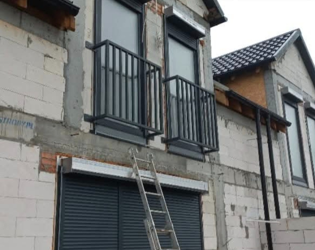Surowe balkony z czarnymi balustradami na tle budynku w trakcie budowy, z widoczną drabiną. Montaż rolet okiennych.