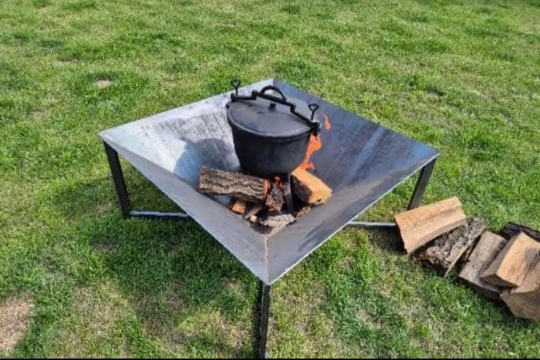 Metalowy kosz na ognisko w kształcie trapezu, z paleniskiem wypełnionym drewnem i czarnym garnkiem. Kosz stoi na trawie, obok leżą kawałki drewna.