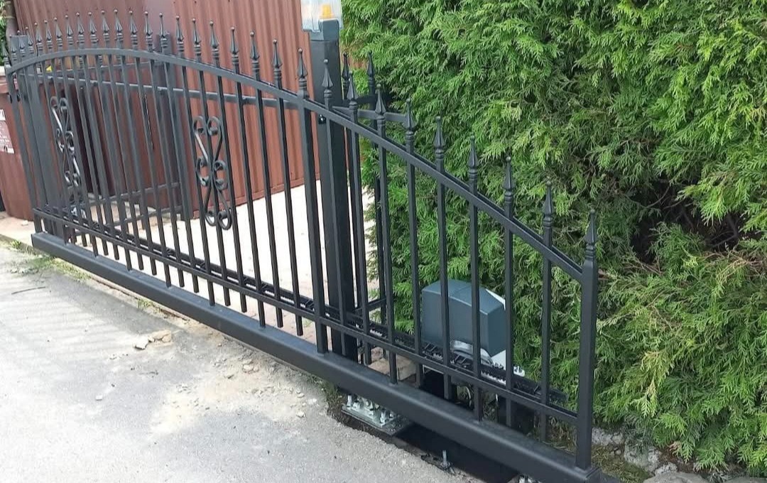Czarna, przesuwna brama wjazdowa z automatyką, zdobiona delikatnym wzorem, na tle zielonego żywopłotu i brązowej ściany. Widoczny mechanizm napędowy.
