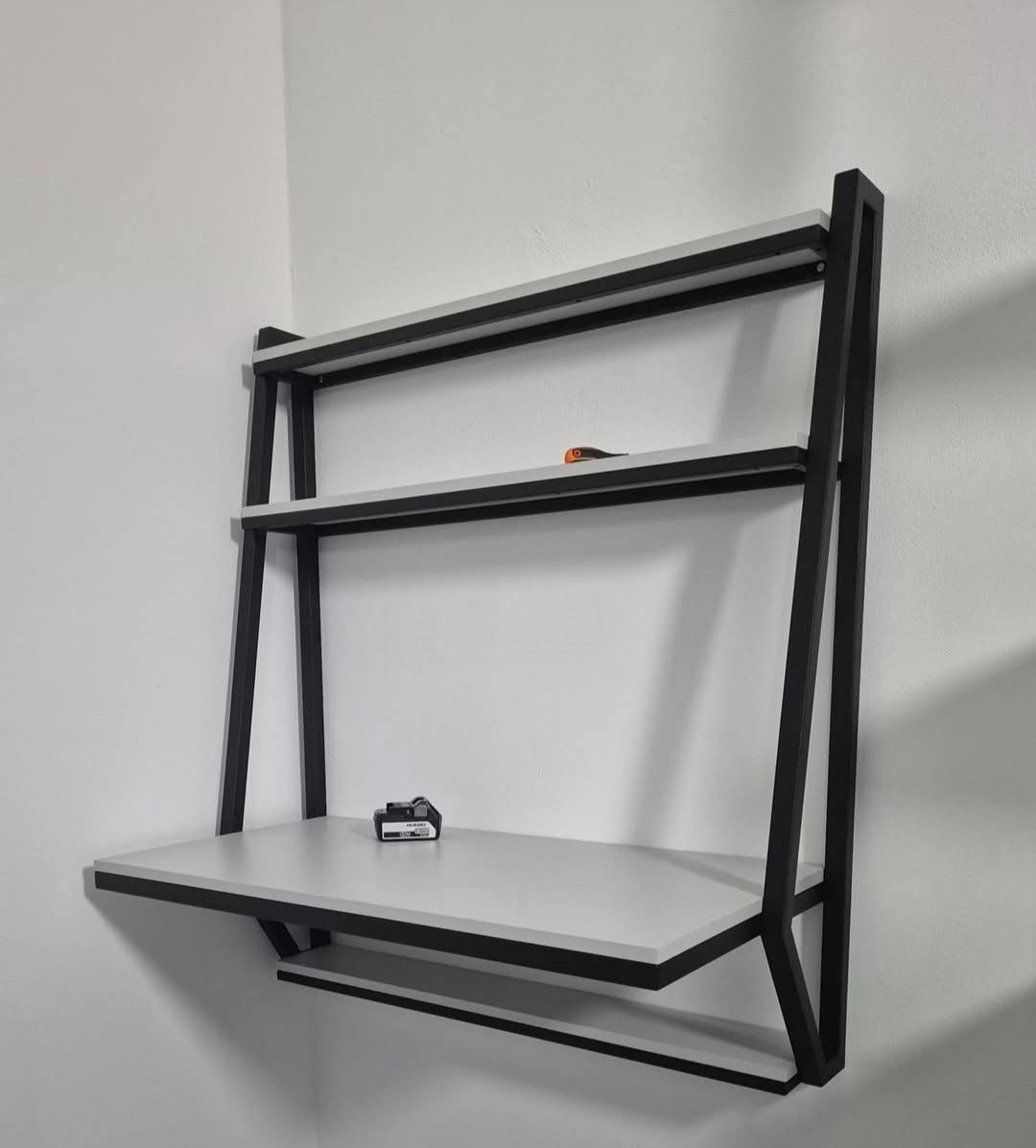 Stylowy, metalowy regał z półkami, wykonany w Gnieznie. Czarna rama i jasne półki, minimalistyczny design, widok z boku, na półce leży bateria do elektronarzędzi.