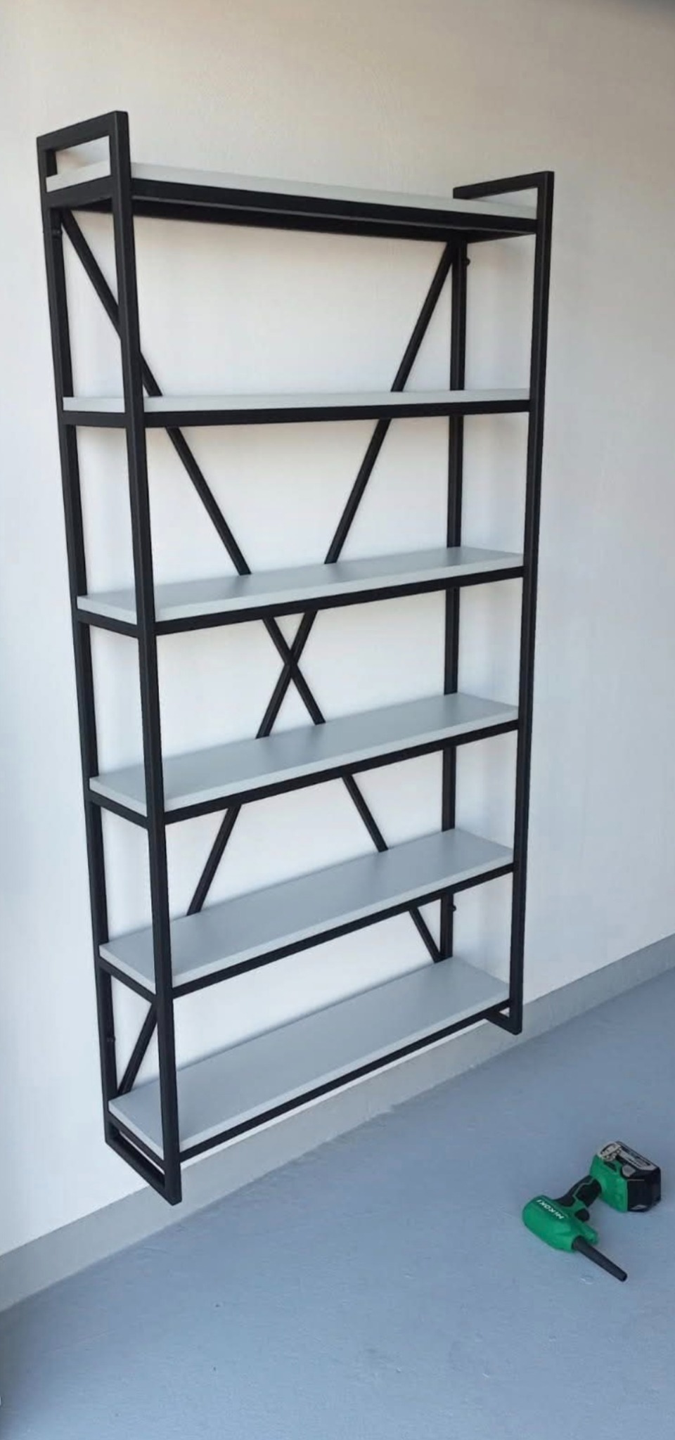 Metalowy regał z jasnymi półkami, czarna konstrukcja z ukośnymi wzmocnieniami. Na podłodze wiertarko-wkrętarka. Minimalistyczny design, nowoczesny styl.