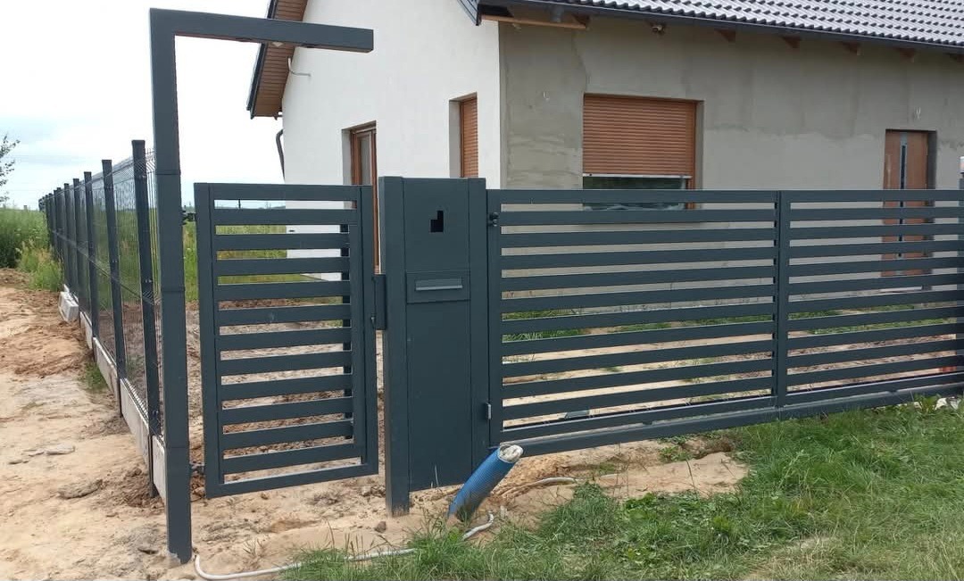 Nowoczesna, pozioma brama wjazdowa i ogrodzenie panelowe w kolorze antracytowym. Brama uchylona, widoczny fragment domu w tle. Słupek oświetleniowy.
