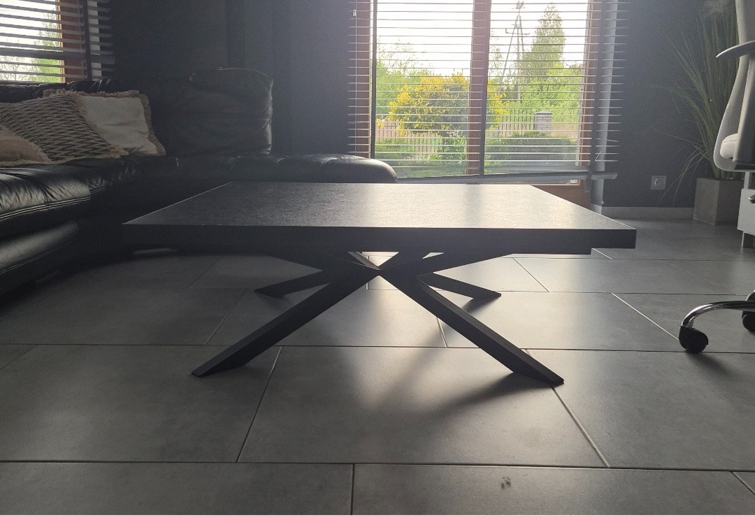 Stylowy, nowoczesny stolik kawowy z ciemnym blatem i geometryczną, metalową podstawą na tle salonu z dużym oknem i skórzaną sofą.