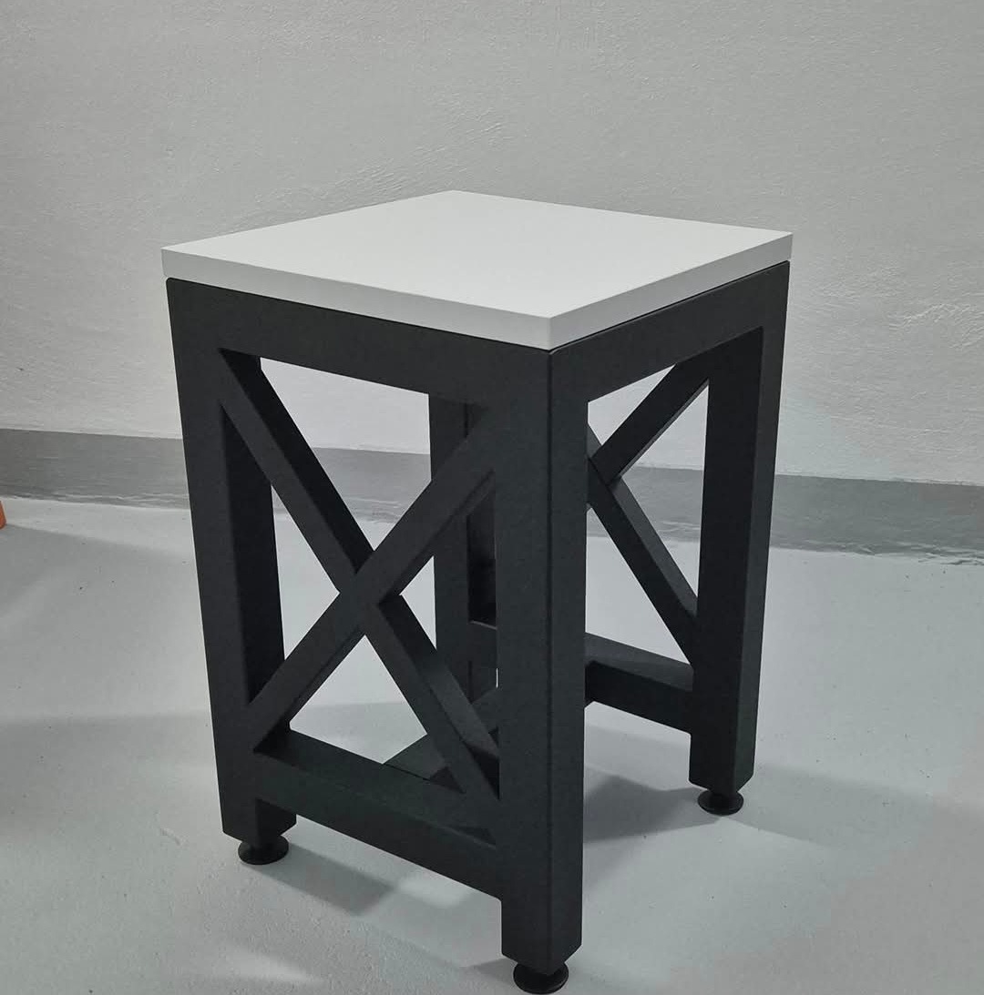 Stolik kawowy z czarną, metalową podstawą w kształcie litery X i białym, kwadratowym blatem, stojący na szarym tle.