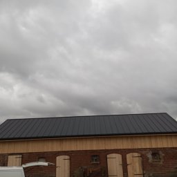 Michał Gałecki Konstrukcje drewniane - Budynek z nowym, ciemnym dachem z blachy trapezowej i drewnianą elewacją. Częściowo widoczny biały bus z otwartym bagażnikiem. Pochmurne niebo w tle.