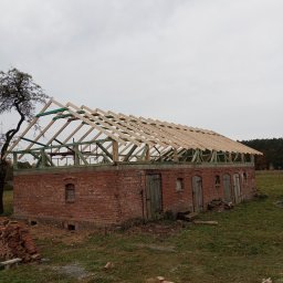 Michał Gałecki Konstrukcje drewniane - Ceglany budynek w trakcie budowy dachu z drewnianą konstrukcją więźby. Widoczne drewniane elementy i ceglane ściany. Na pierwszym planie leżą pnie drzew.