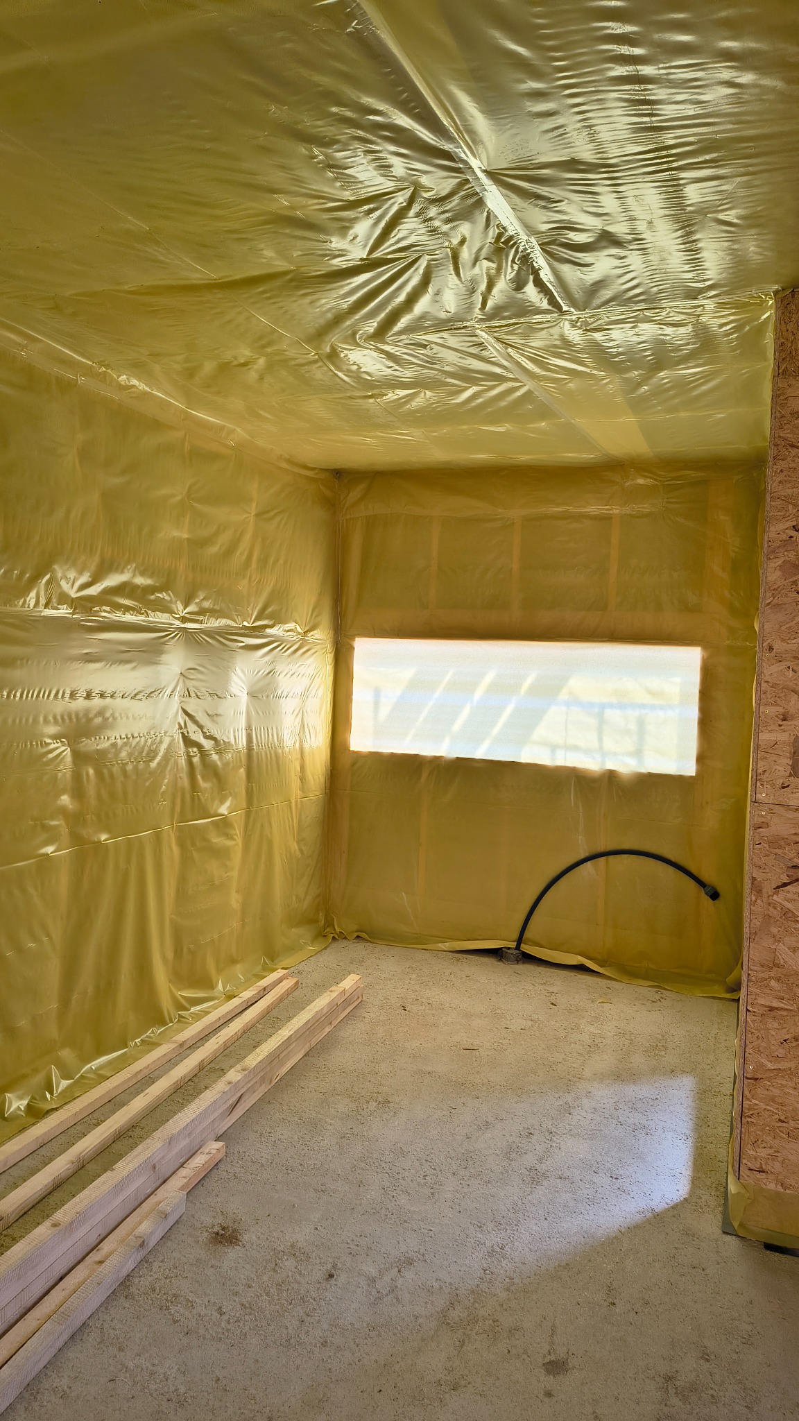 Wnętrze w trakcie budowy domu szkieletowego: ściany i sufit pokryte żółtą folią izolacyjną, widoczne drewniane elementy konstrukcyjne i fragment okna. Na podłodze leżą drewniane belki.