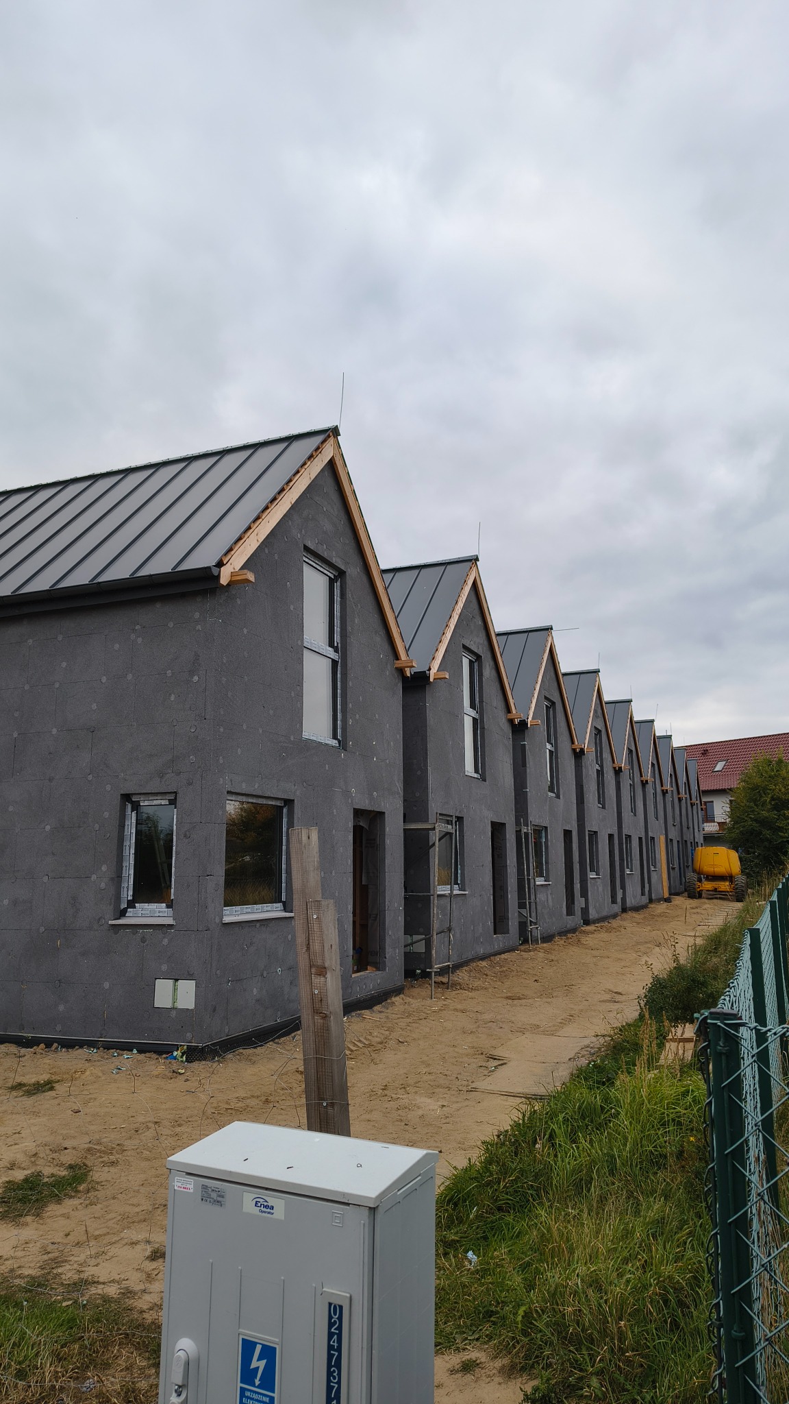Szereg identycznych, surowych domów szkieletowych z szarą elewacją i ciemnymi dachami, w trakcie budowy, z widocznymi oknami i elementami konstrukcyjnymi.