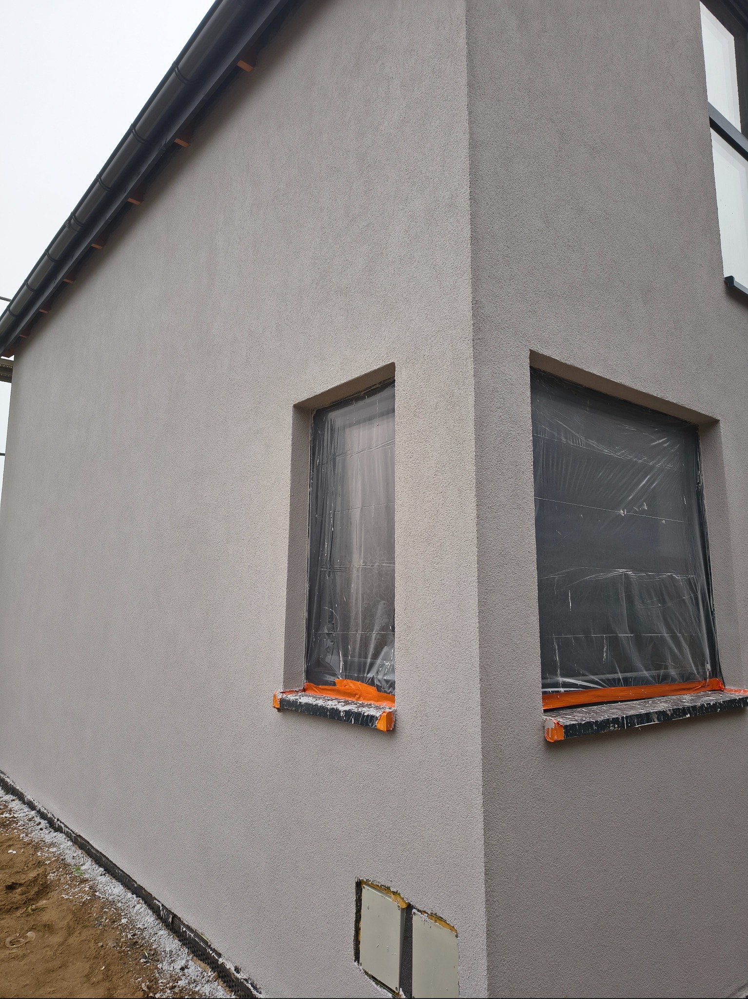 Fragment elewacji budynku z oknami zabezpieczonymi folią malarską. Widoczna faktura tynku i fragment dachu z rynną. Ujęcie z dołu, podkreślające bryłę budynku.