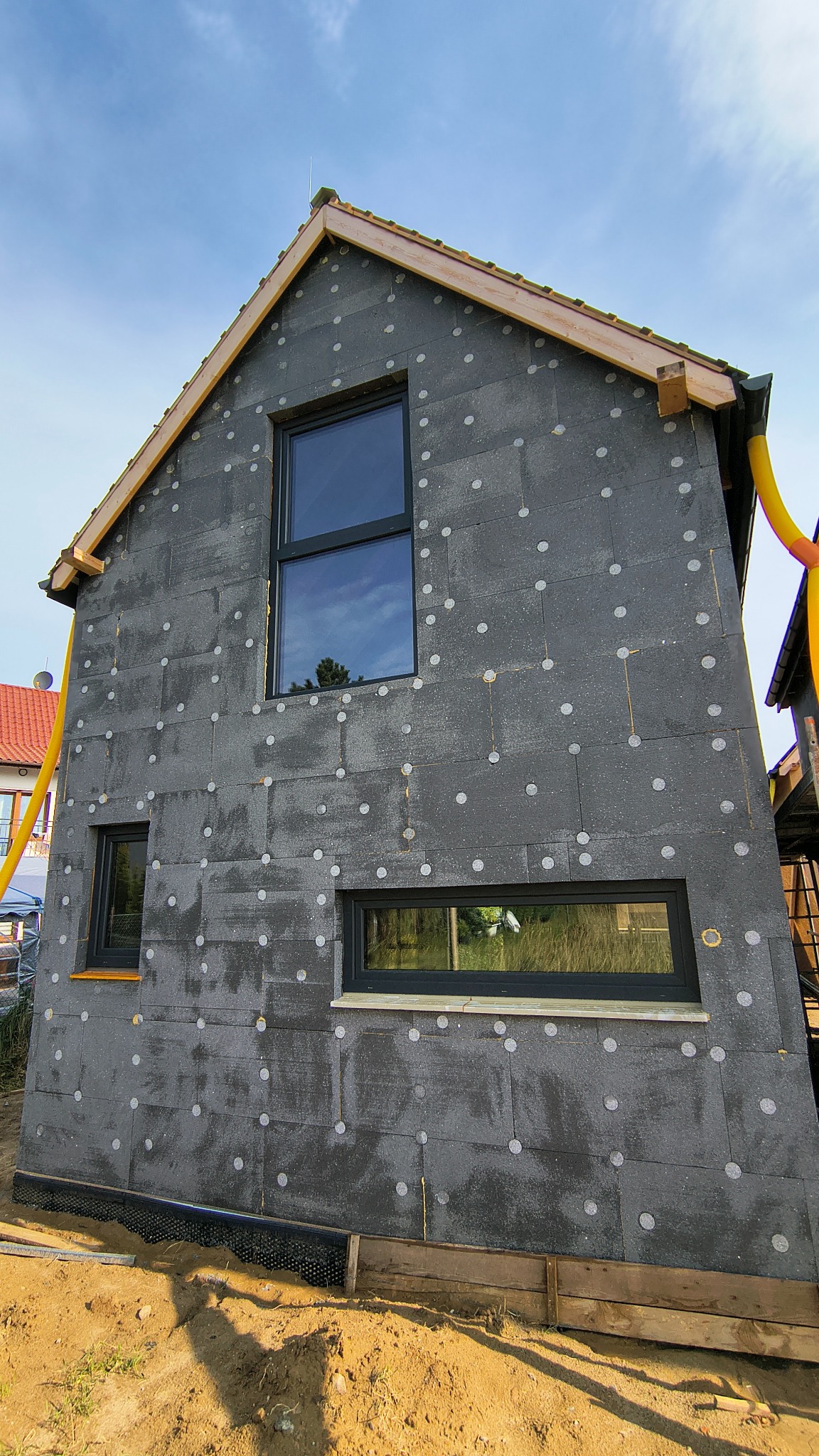 Budowa domu szkieletowego: ściana z oknami, pokryta szarą izolacją z białymi kołkami, widoczne elementy dachu i rynny. Ujęcie z dołu, błękitne niebo w tle.