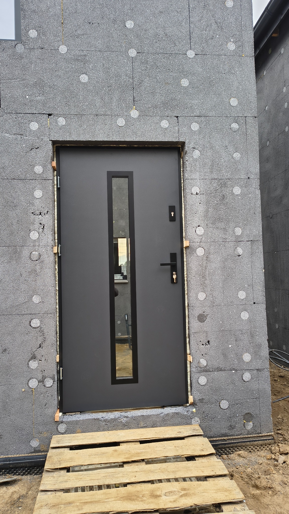 Szare drzwi wejściowe z wąskim przeszkleniem, zamontowane w ścianie z szarego styropianu grafitowego. Paleta drewniana ułożona przed wejściem.