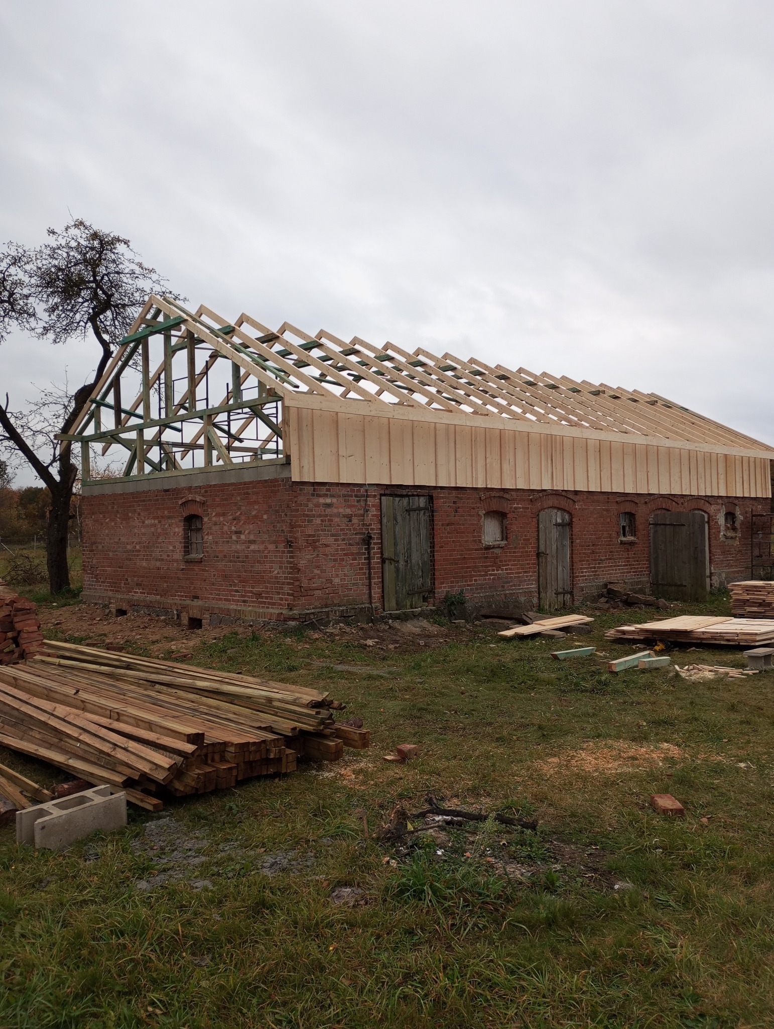 Ceglany budynek z drewnianą konstrukcją dachu w trakcie budowy. Widoczne deski i elementy więźby. Na pierwszym planie stos drewna. Pochmurne niebo w tle.