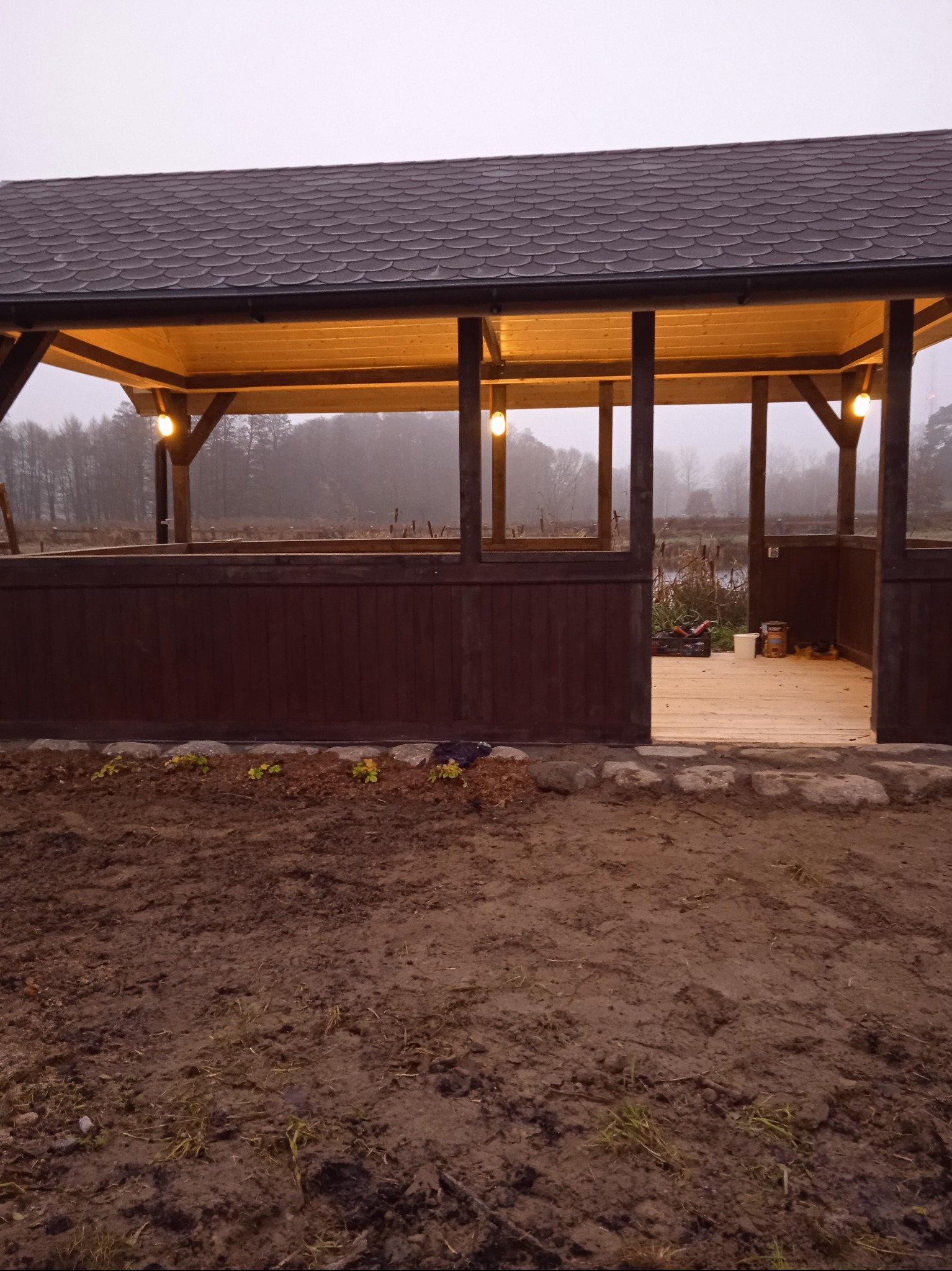 Drewniana altana z oświetleniem w ogrodzie, widok z zewnątrz na drewnianą konstrukcję z dachem pokrytym gontem bitumicznym. W tle zarośla i mgła.