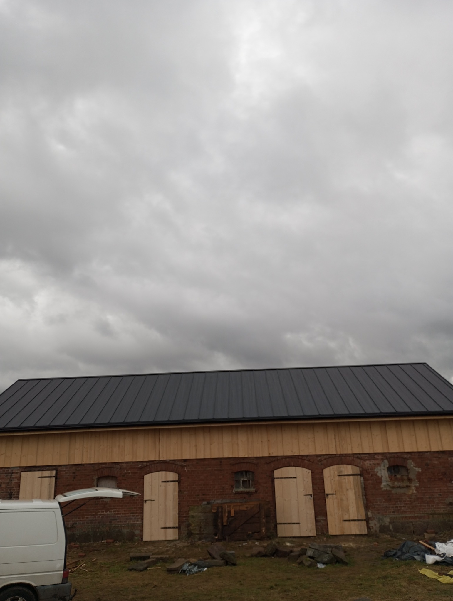 Budynek z nowym, ciemnym dachem z blachy trapezowej i drewnianą elewacją. Częściowo widoczny biały bus z otwartym bagażnikiem. Pochmurne niebo w tle.