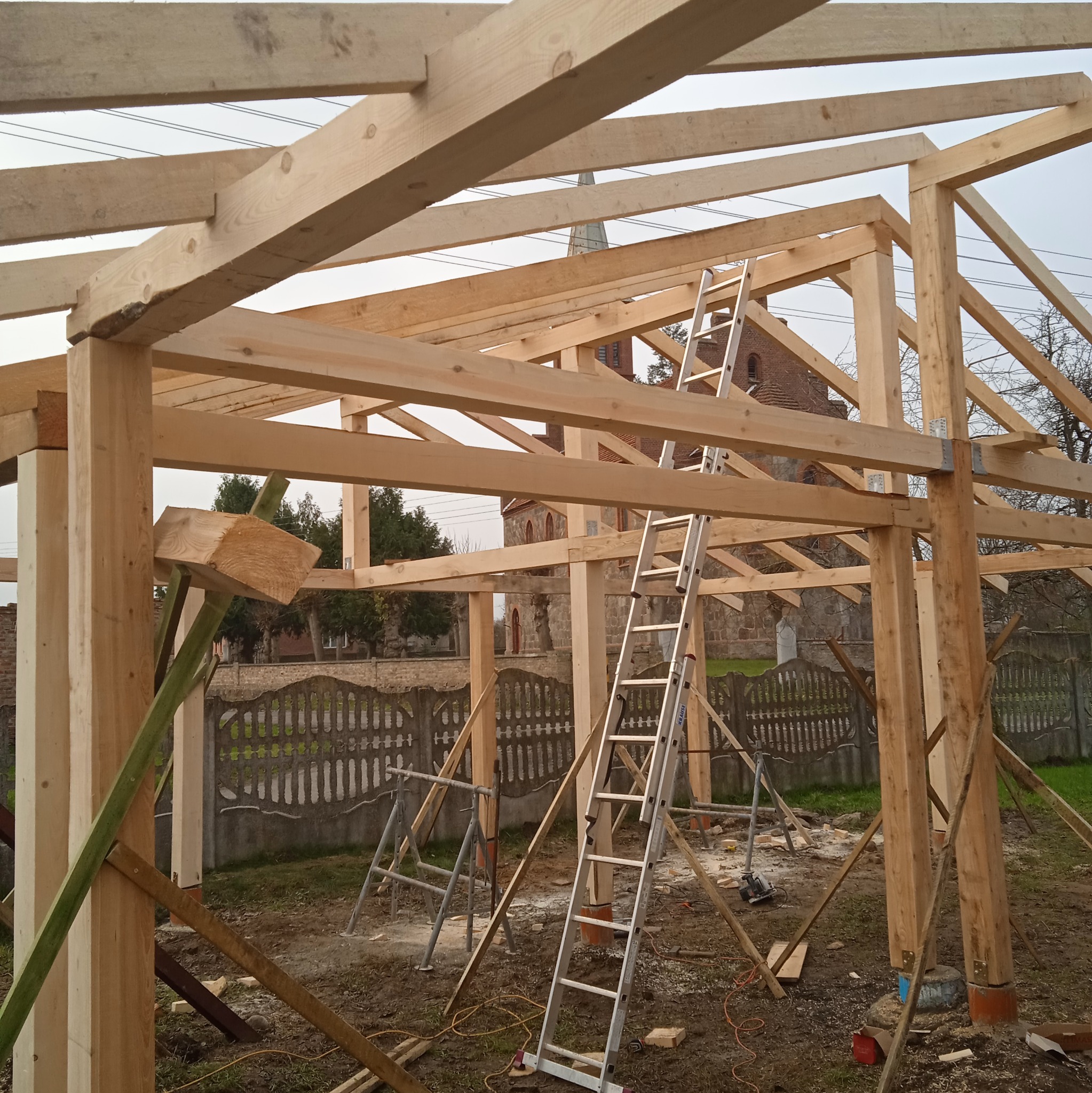 Wznoszenie drewnianej konstrukcji szkieletowej z widoczną więźbą dachową i podporami tymczasowymi. W tle ceglany budynek kościoła.