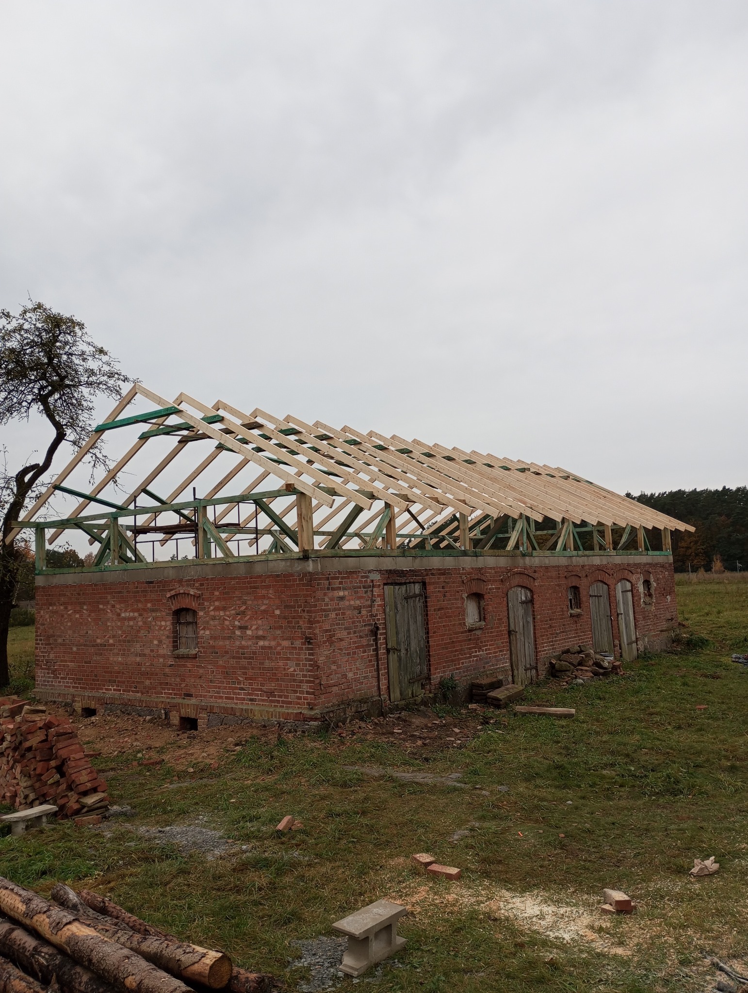 Ceglany budynek w trakcie budowy dachu z drewnianą konstrukcją więźby. Widoczne drewniane elementy i ceglane ściany. Na pierwszym planie leżą pnie drzew.
