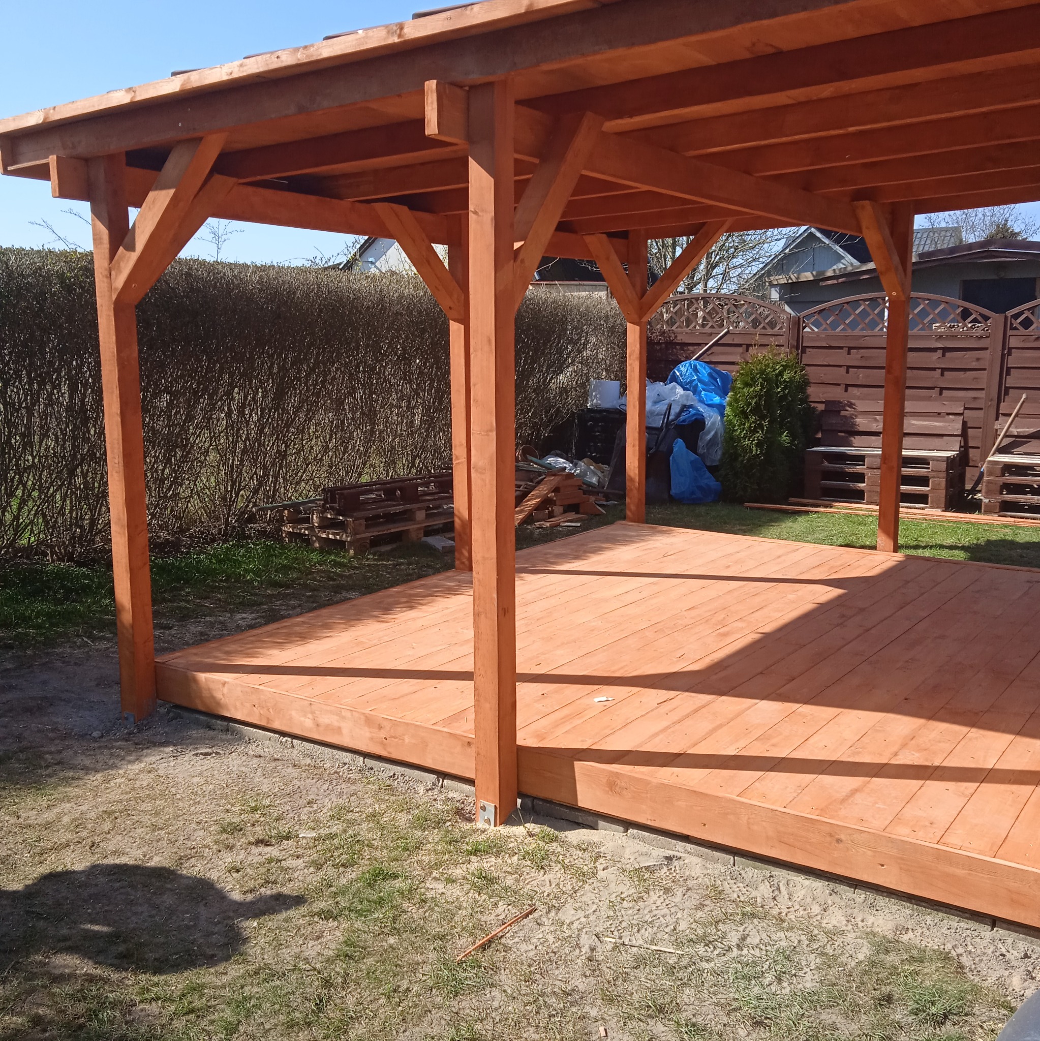 Nowa, drewniana pergola z podłogą z desek, w ogrodzie. Widoczne elementy konstrukcyjne i podłoże. Jasne, naturalne światło.
