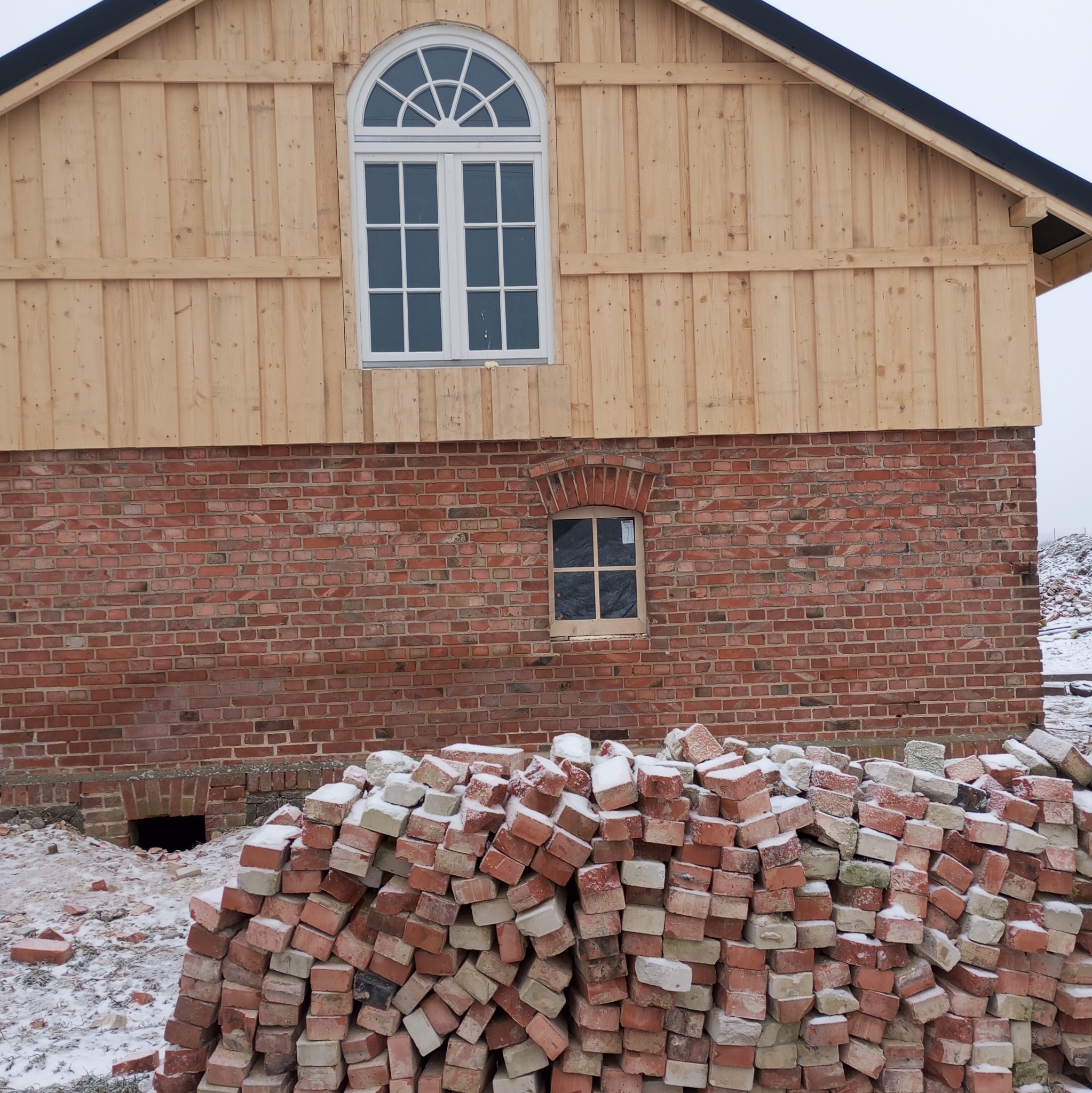 Budynek w trakcie budowy: ceglana ściana z małym oknem i drewniana konstrukcja z dużym oknem łukowym. Na pierwszym planie sterta cegieł pokryta śniegiem.