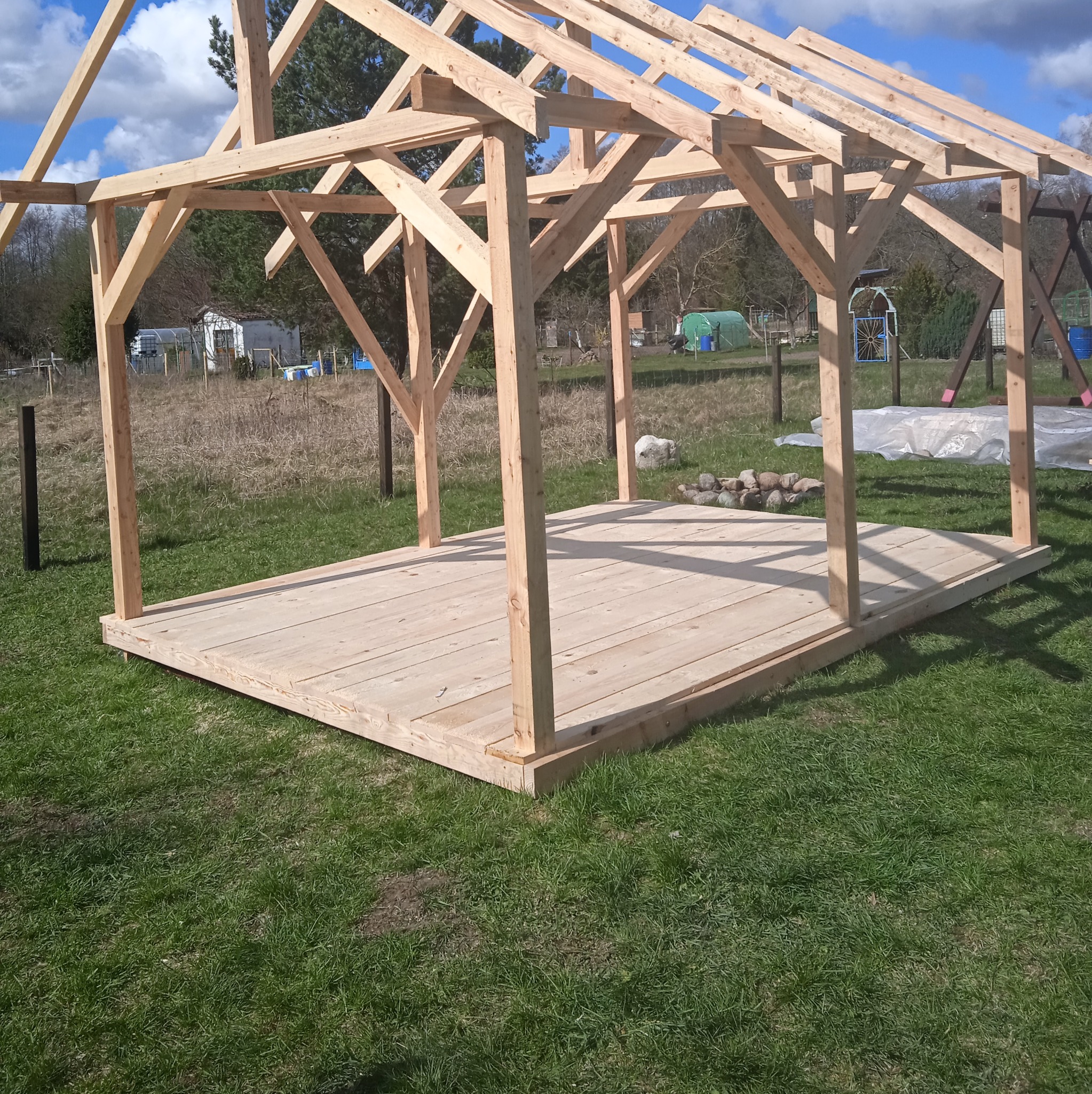 Surowa drewniana konstrukcja altany w ogrodzie, widoczne słupy, podłoga i szkielet dachu, w tle zieleń i błękitne niebo z chmurami. Etap budowy.
