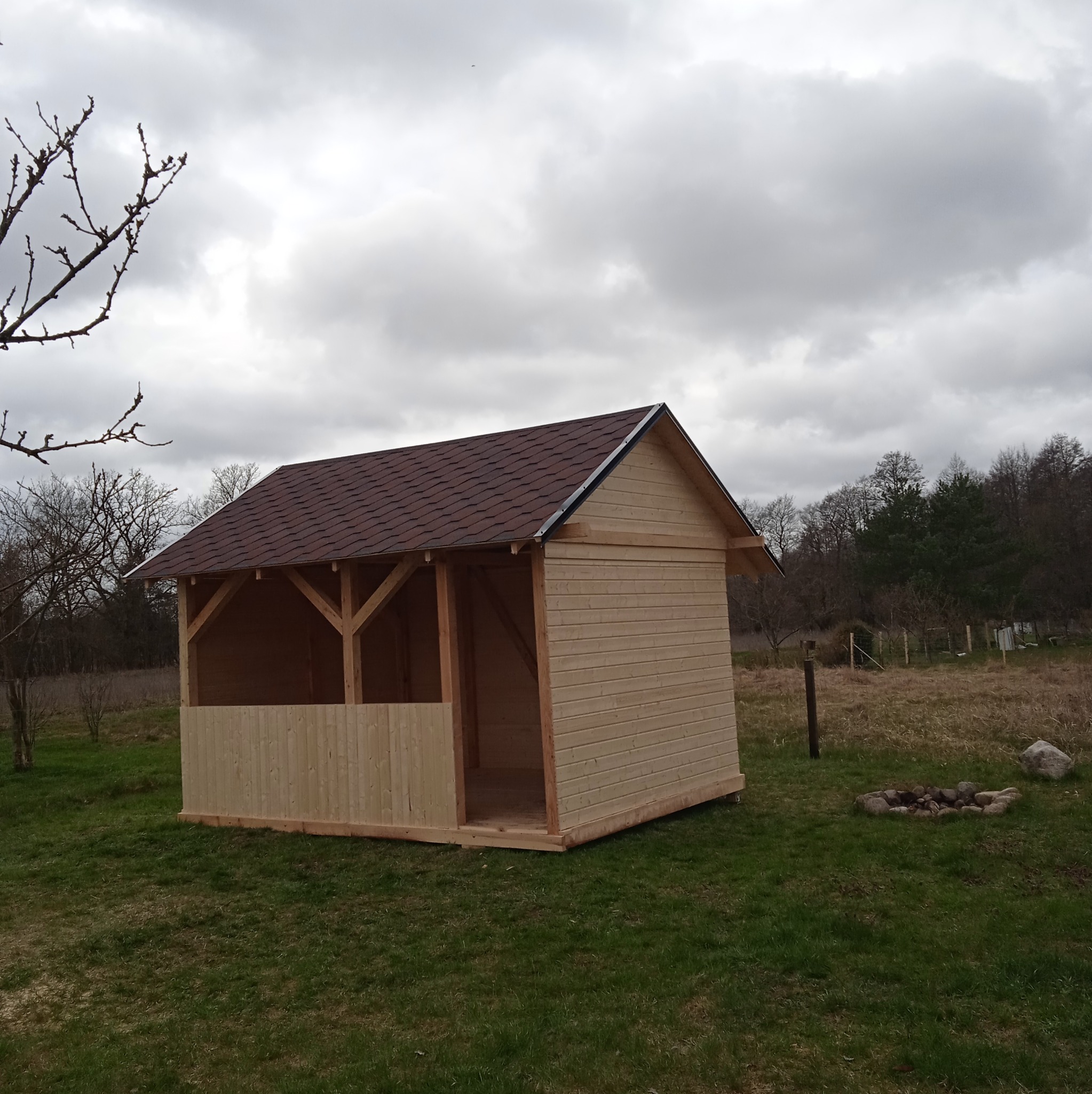 Drewniana altana z brązowym dachem z gontu bitumicznego, częściowo osłonięta, na trawiastym terenie w pochmurny dzień. Widoczny fragment drzewa i palenisko.