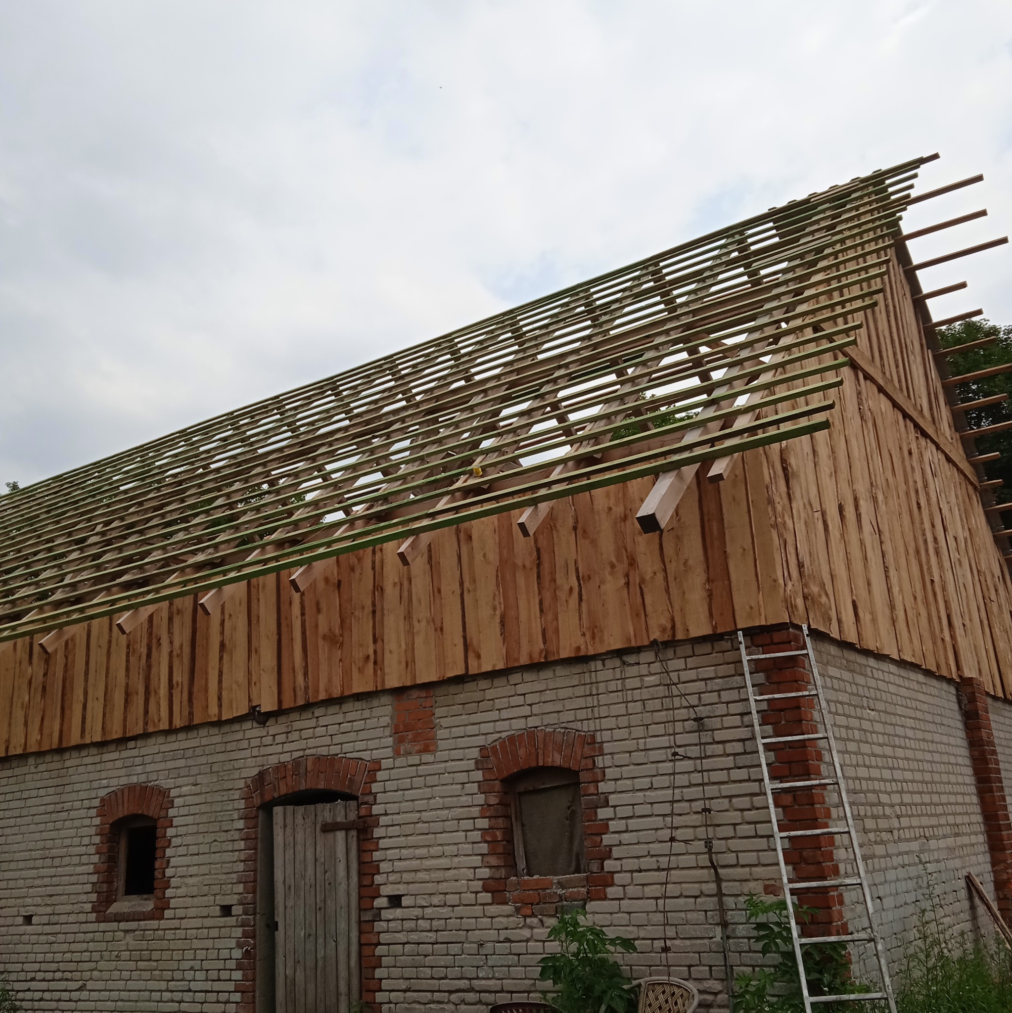 Budynek z cegły i drewna w trakcie budowy dachu. Widoczna więźba dachowa i drewniane deski. Na ścianie oparta drabina. Pochmurne niebo w tle.