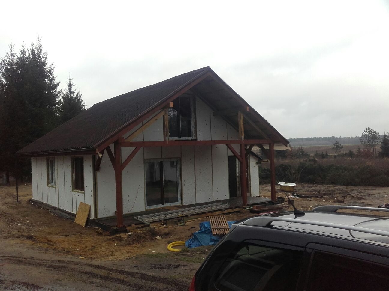 Surowy stan domu szkieletowego z brązowym dachem, widoczne drewniane elementy konstrukcyjne i okna. W tle las i pochmurne niebo. Na pierwszym planie fragment samochodu.