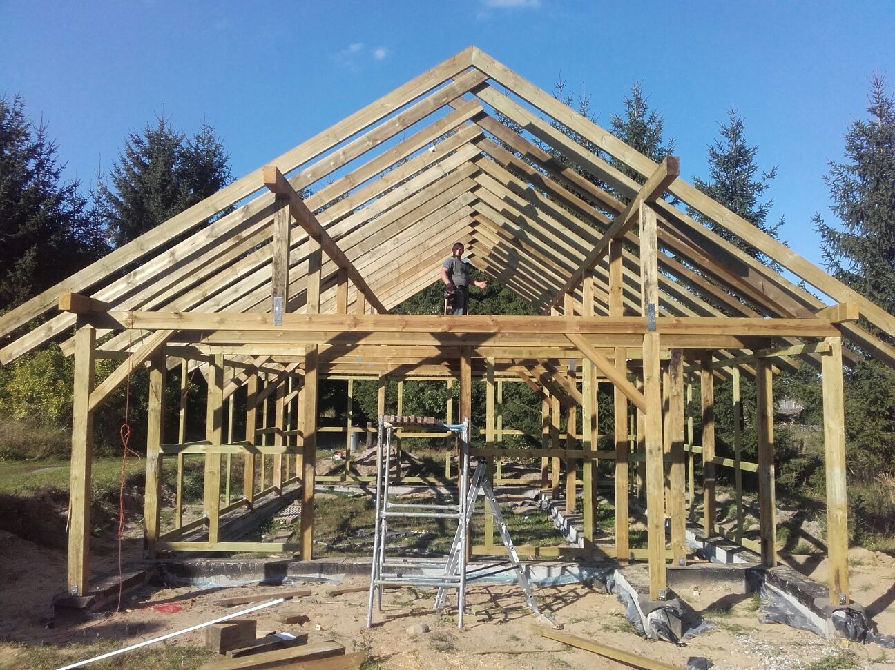 Konstrukcja drewniana domu w trakcie budowy, widoczna więźba dachowa i szkielet ścian. Na górze konstrukcji stoi osoba. W tle drzewa i błękitne niebo.
