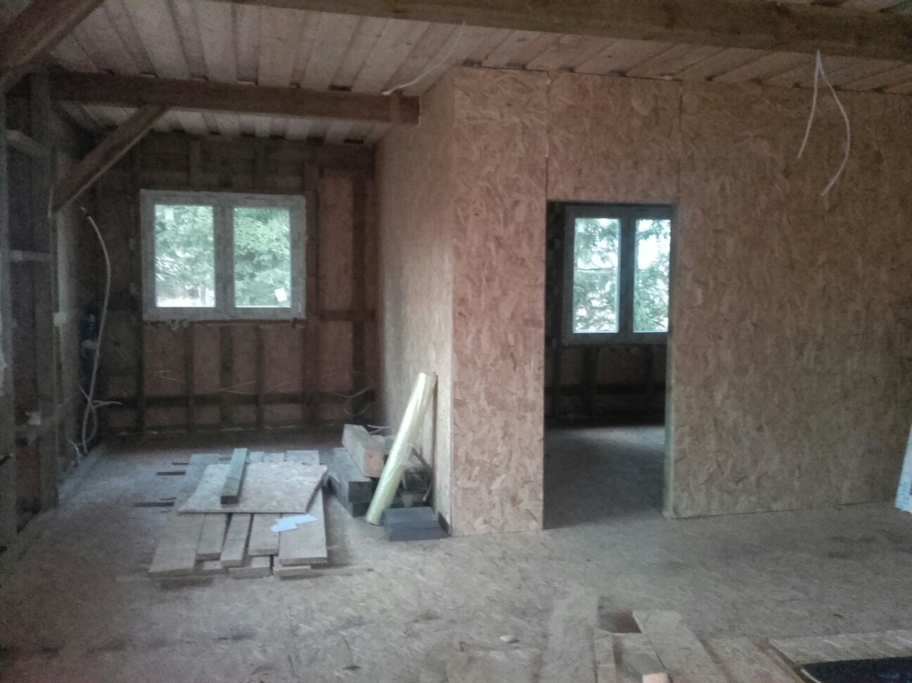 Wnętrze w trakcie budowy: ściany z płyt OSB, drewniana konstrukcja stropu, okna z widokiem na zieleń. Na podłodze leżą deski i materiały budowlane.