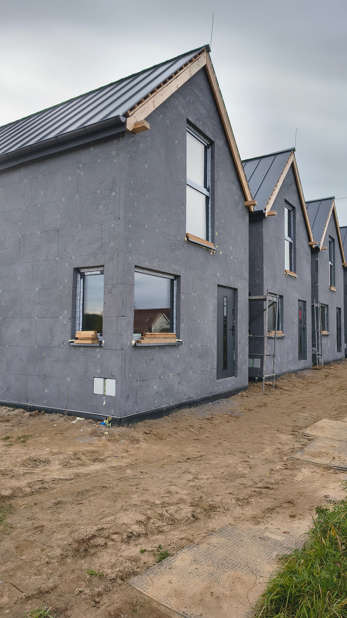 Surowe domy szkieletowe w trakcie budowy, z widoczną izolacją zewnętrzną i drewnianą konstrukcją dachu, ustawione szeregowo na piaszczystym terenie.