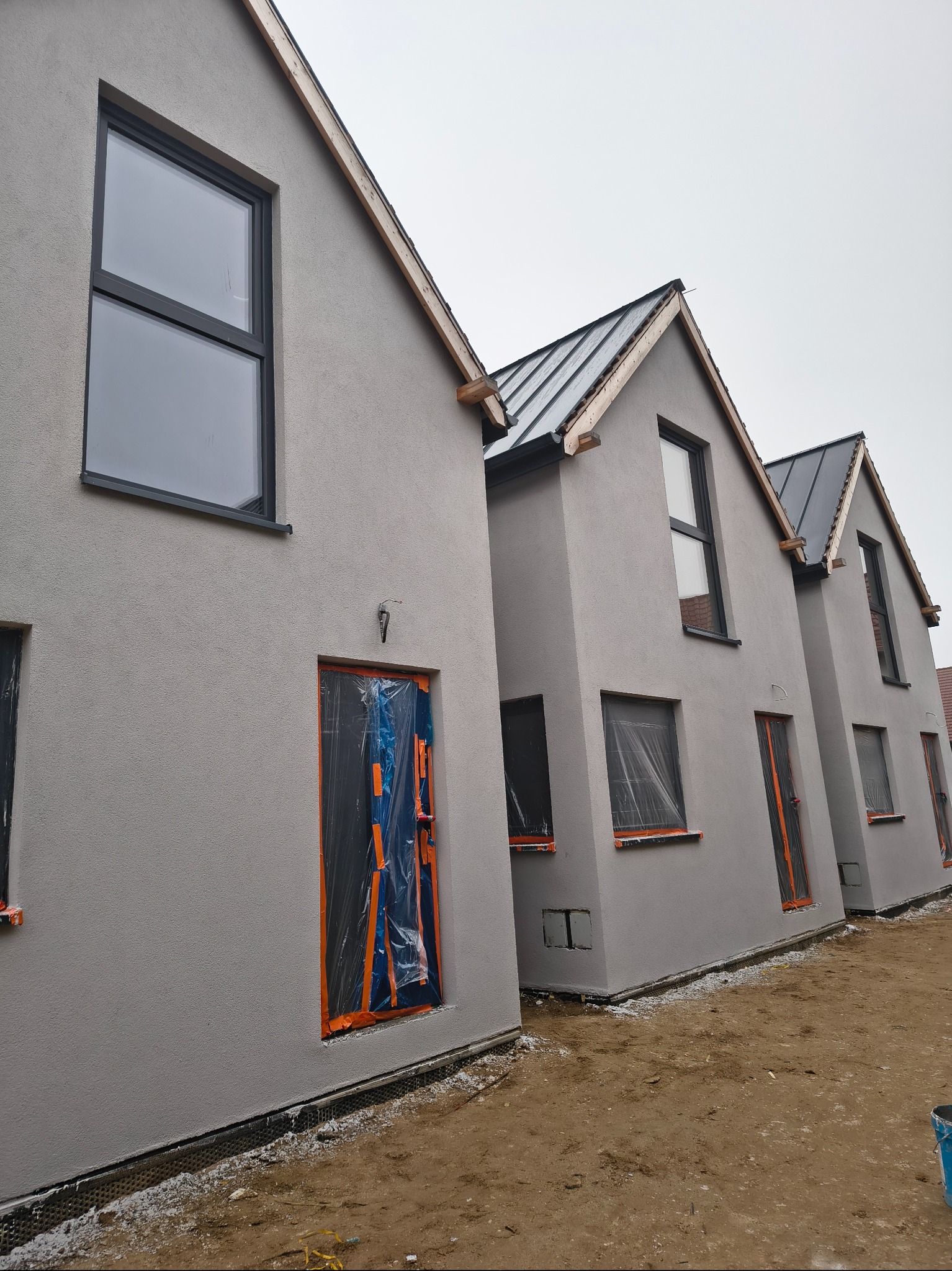 Trzy identyczne, szare domy w zabudowie szeregowej z czarnymi oknami i dachem z blachy. Okna i drzwi zabezpieczone folią. Widok z zewnątrz, w trakcie budowy.