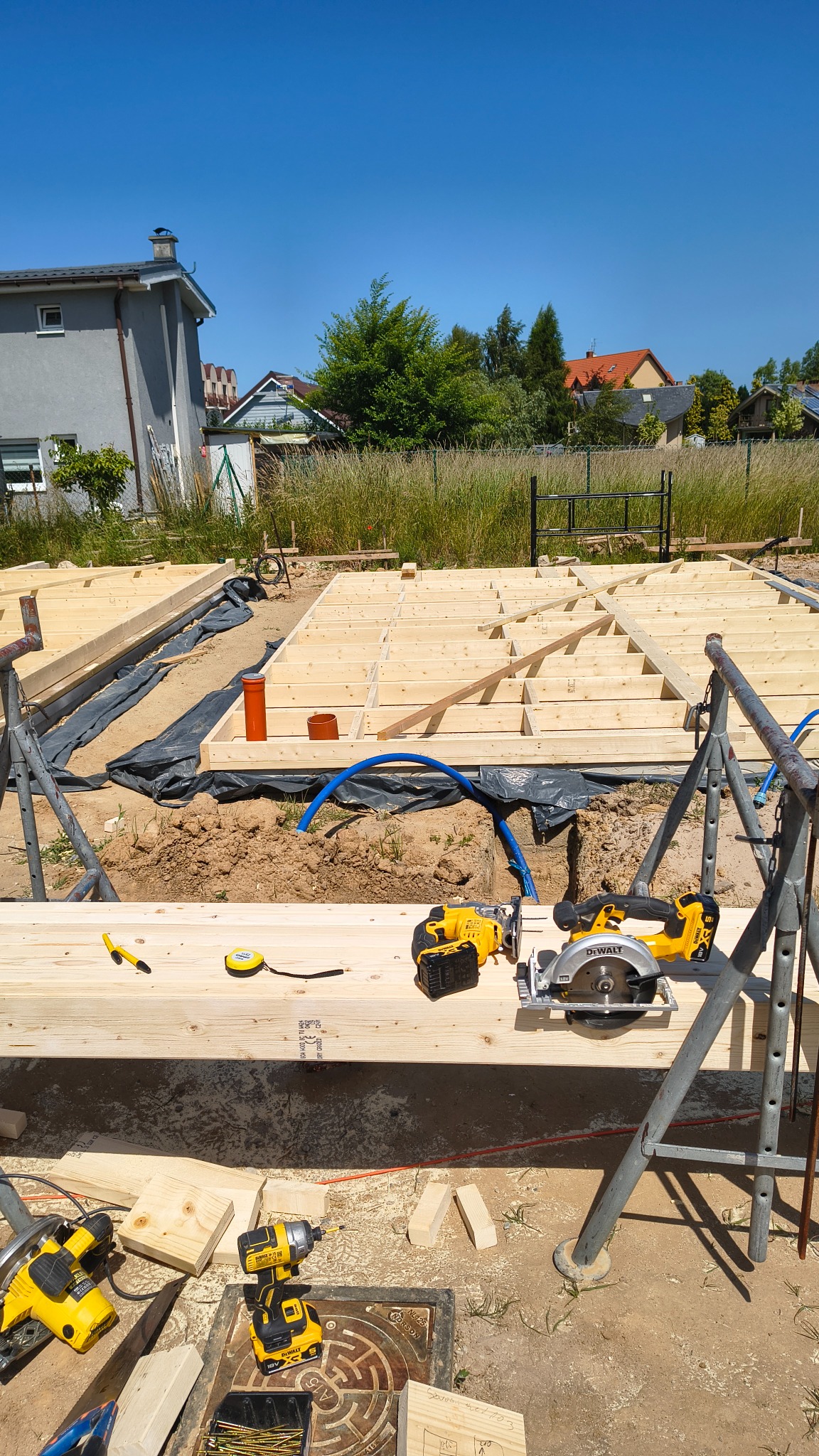 Budowa domu szkieletowego: drewniana konstrukcja fundamentów z narzędziami DeWalt na pierwszym planie, w tle dom i zieleń w słoneczny dzień.