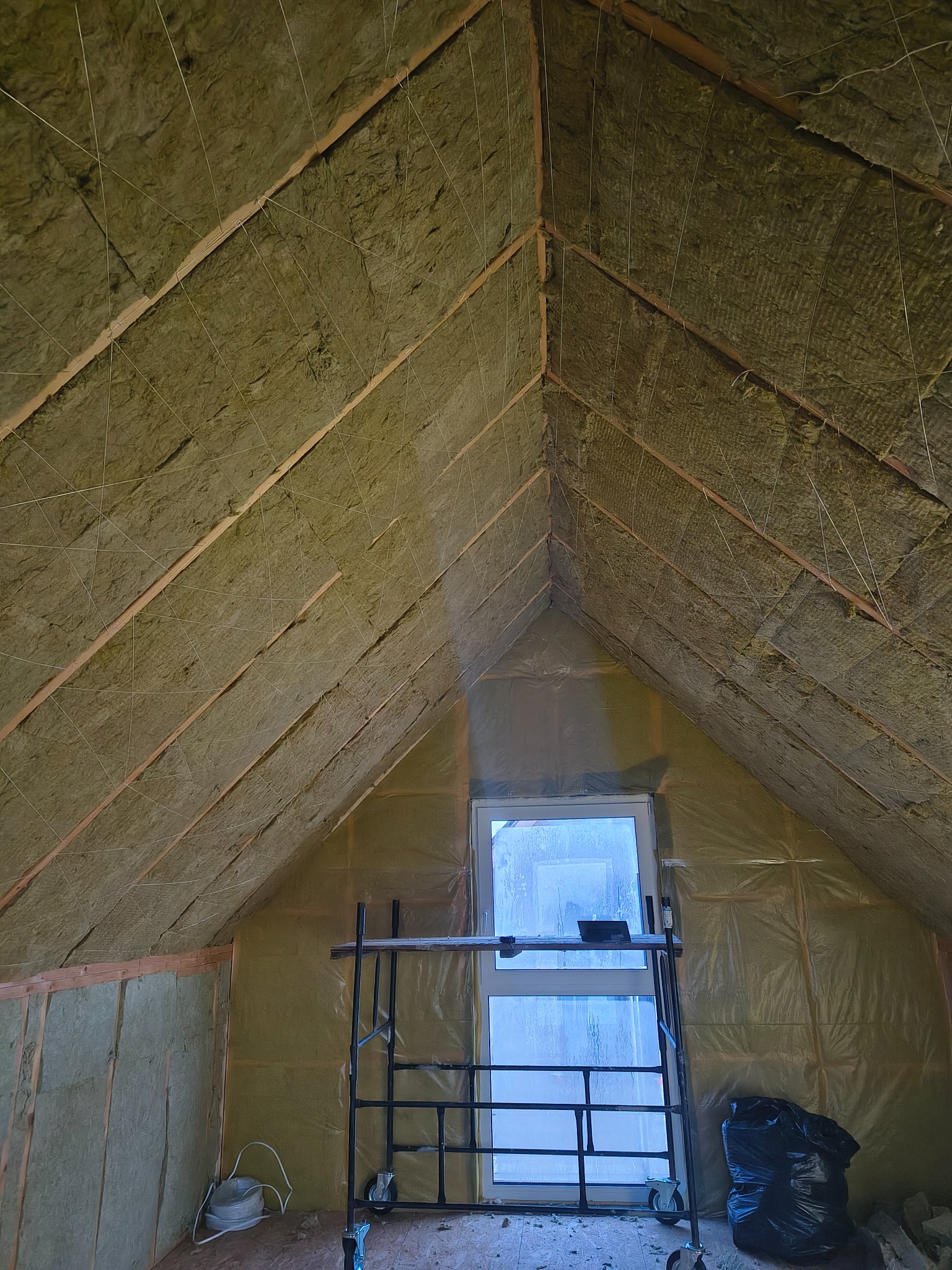 Izolacja poddasza wełną mineralną, widoczne drewniane krokwie i folia paroizolacyjna. U dołu rusztowanie i okno. Prace wykończeniowe na poddaszu.