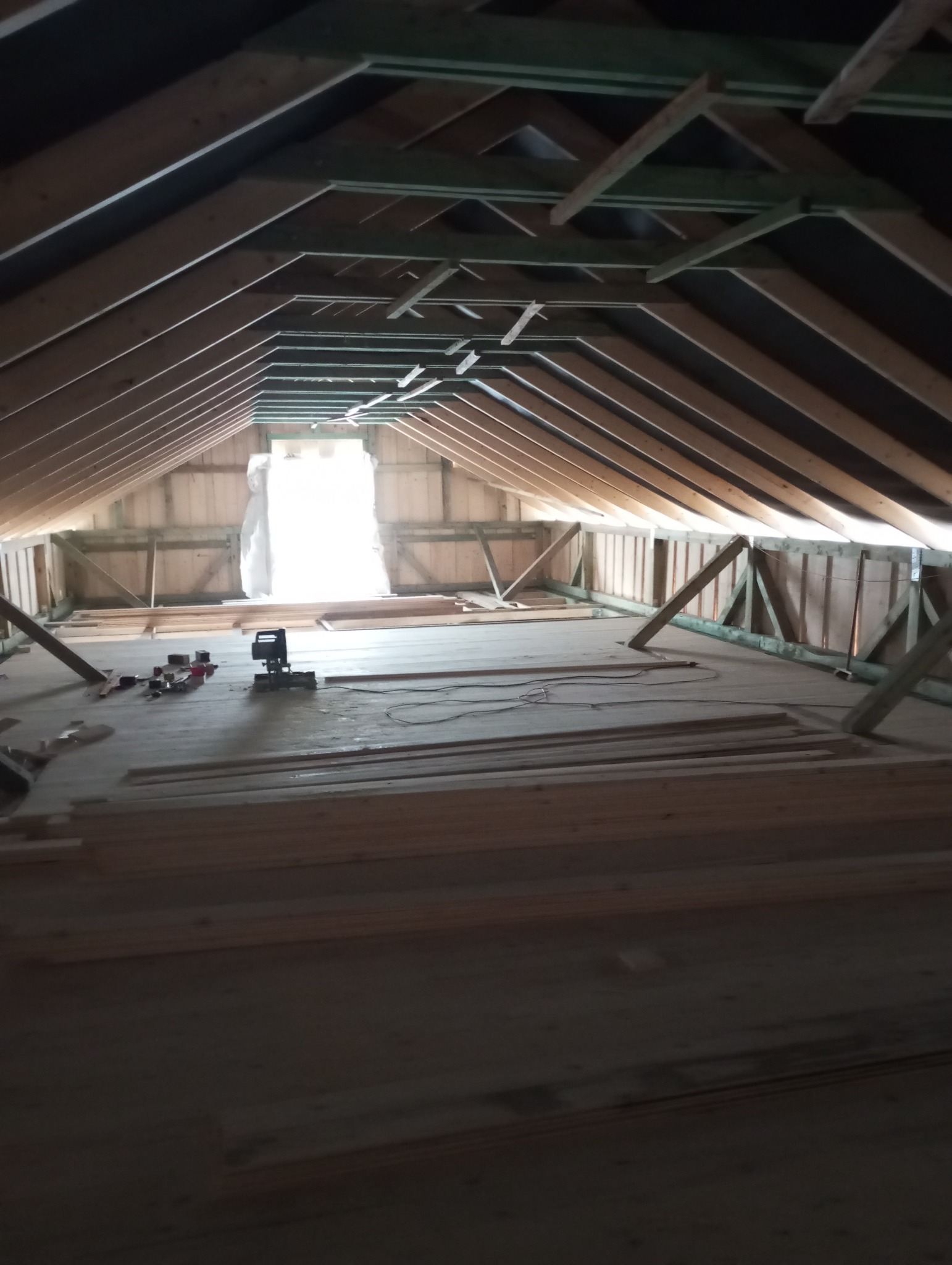 Wnętrze poddasza w trakcie budowy, widoczne drewniane elementy konstrukcyjne dachu i podłogi, narzędzia pracy. Jasne światło wpada przez otwór w ścianie szczytowej.
