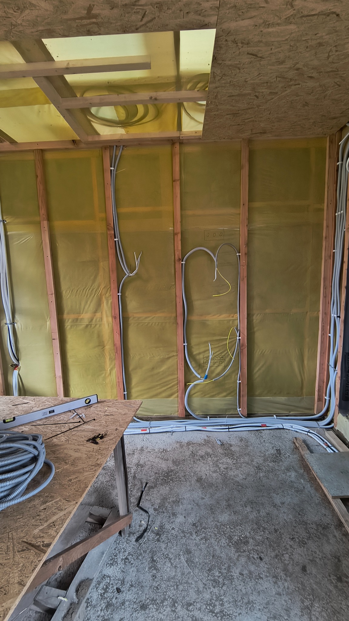 Wnętrze w trakcie budowy: drewniana konstrukcja ścian z izolacją, poprowadzone szare rury instalacji elektrycznej, poziomica na stole roboczym. Widoczne elementy konstrukcyjne.