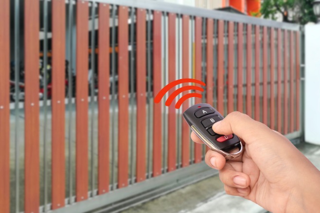 Osoba obsługuje pilot zdalnego sterowania przy drewniano-metalowej bramie posesji, sygnalizując jej otwieranie.
