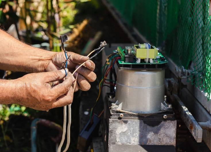 Zbliżenie na ręce elektryka łączącego przewody przy silniku sterującym bramą, widoczne elementy elektroniki i przekładni.