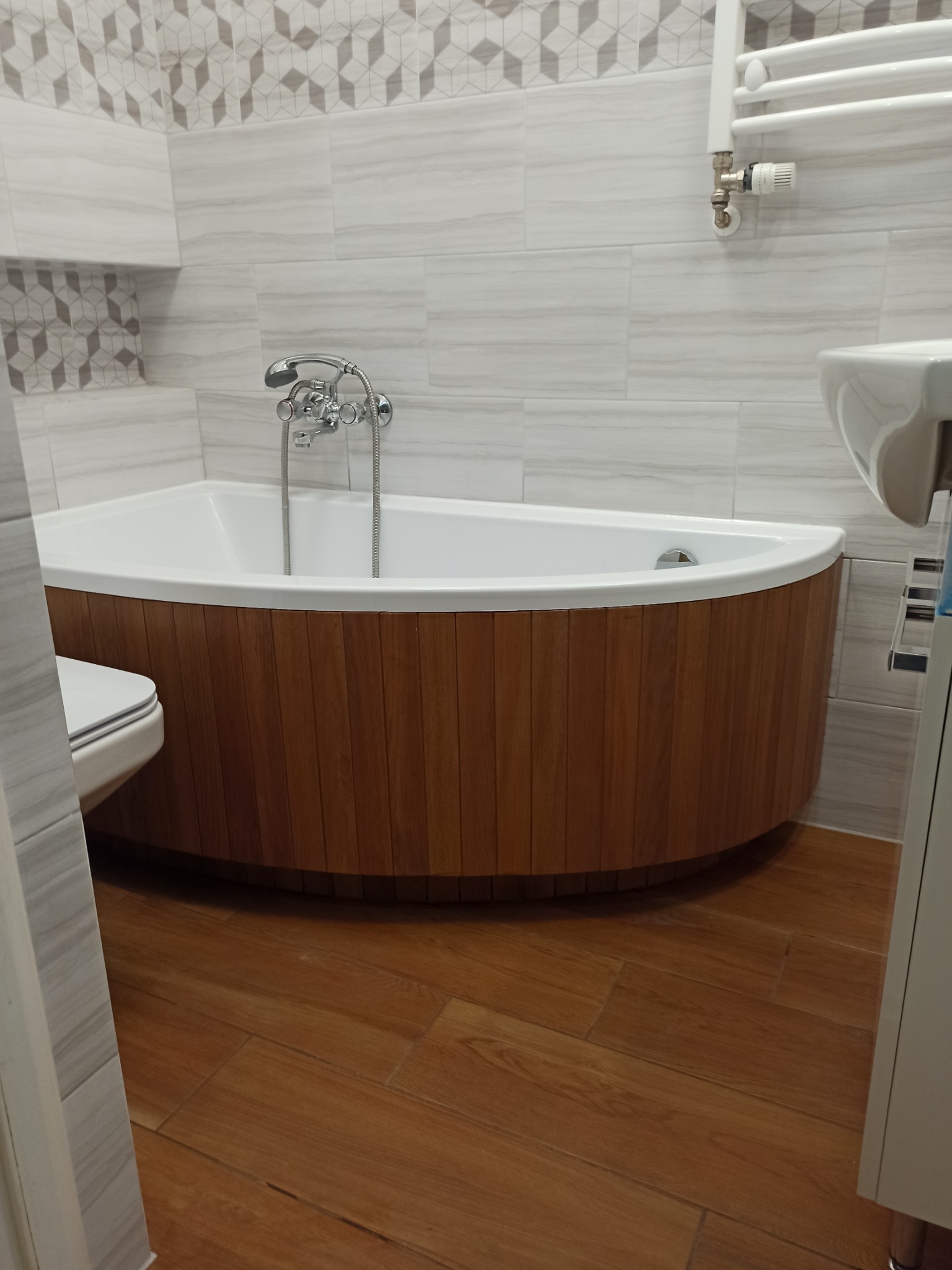 Nowoczesna łazienka po renowacji: narożna wanna z obudową z drewnianych paneli, jasne płytki imitujące marmur z dekoracyjnym pasem mozaiki geometrycznej, drewniana podłoga.