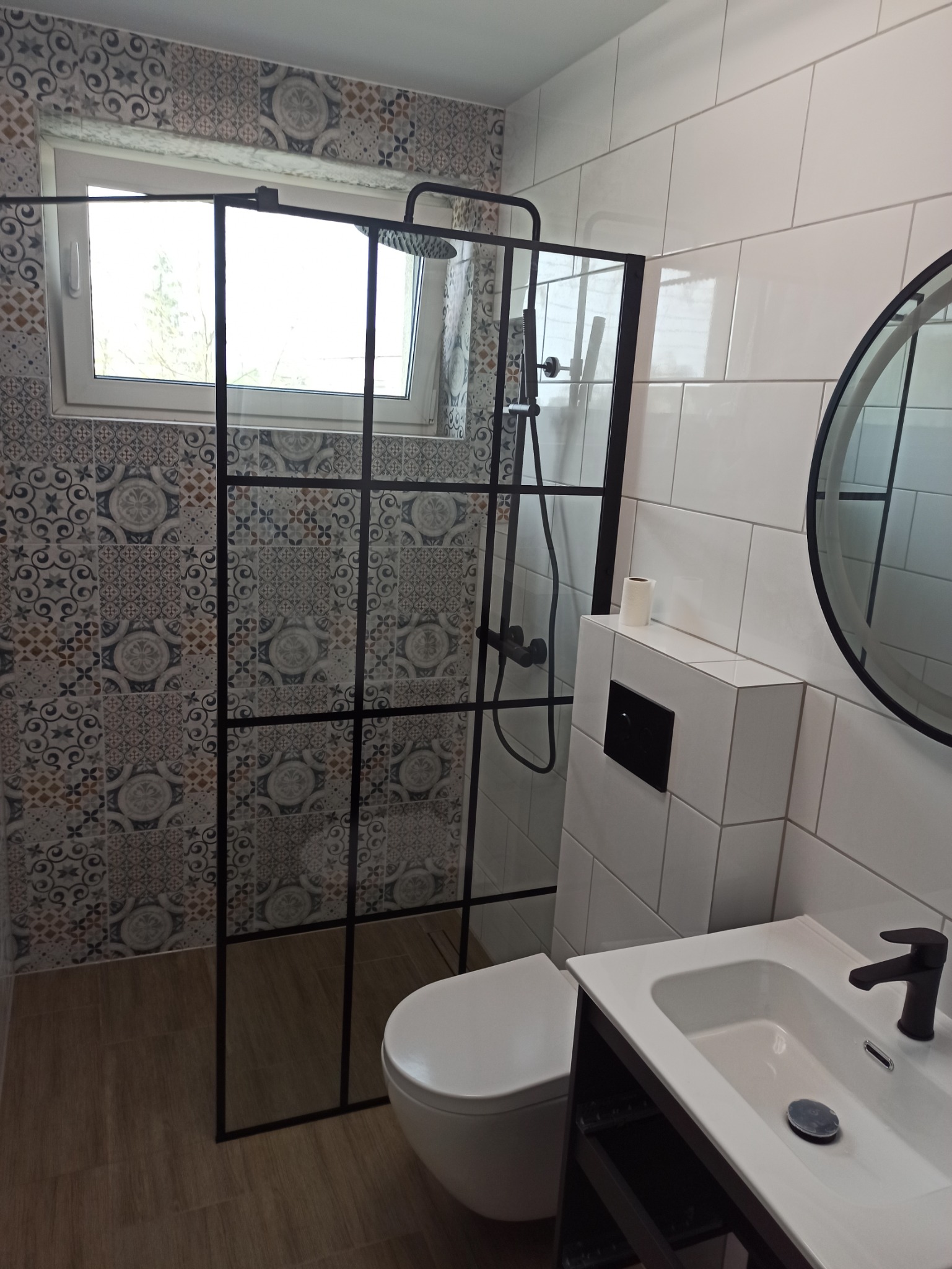 Nowoczesna łazienka po remoncie z kabiną prysznicową typu walk-in z czarnymi szprosami, umywalką na szafce i okrągłym lustrem, wzorzyste płytki pod oknem.