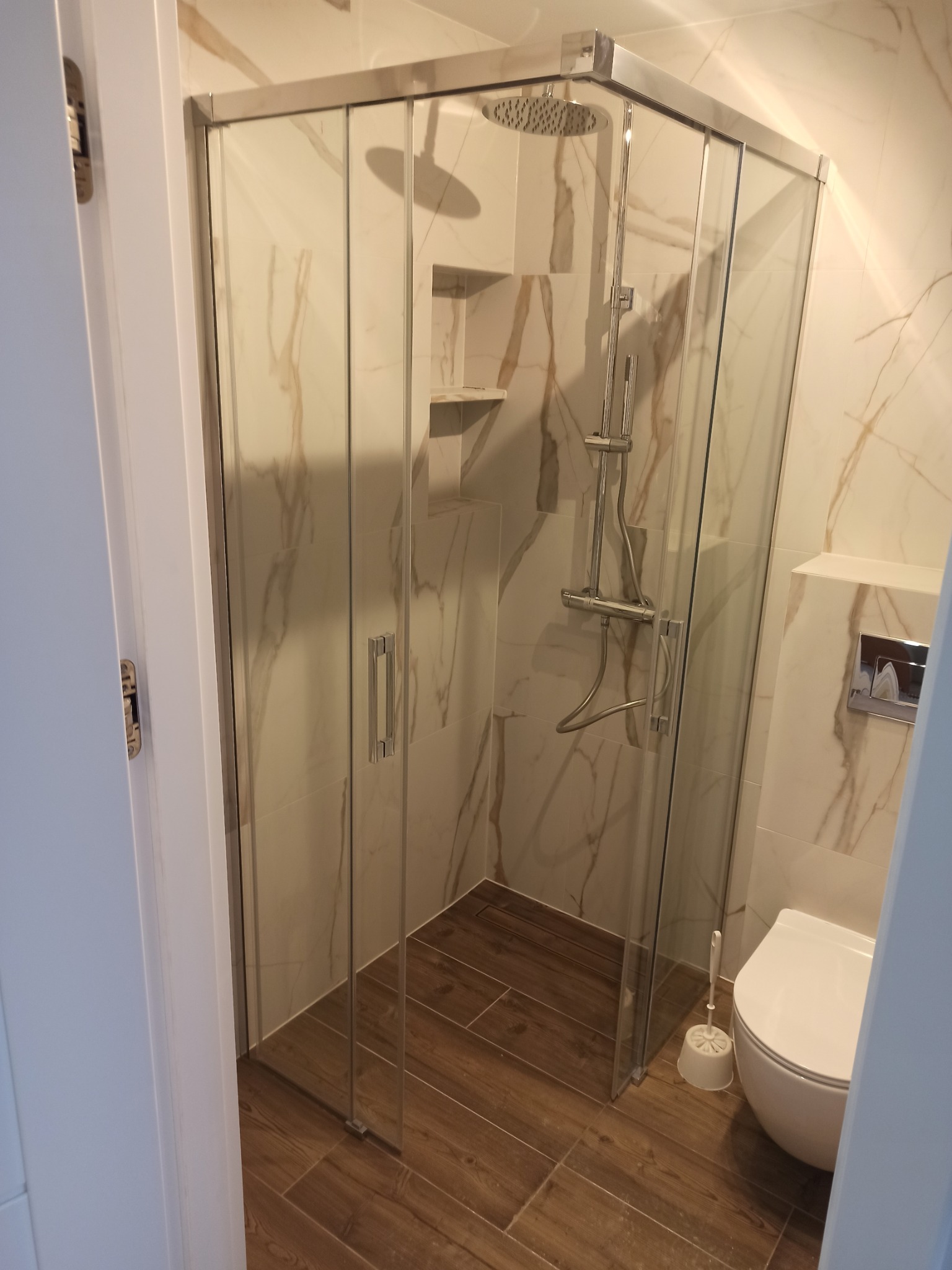 Nowoczesna kabina prysznicowa z deszczownicą w łazience wykończonej płytkami imitującymi marmur i drewnopodobną podłogą, widok zza uchylonych drzwi.