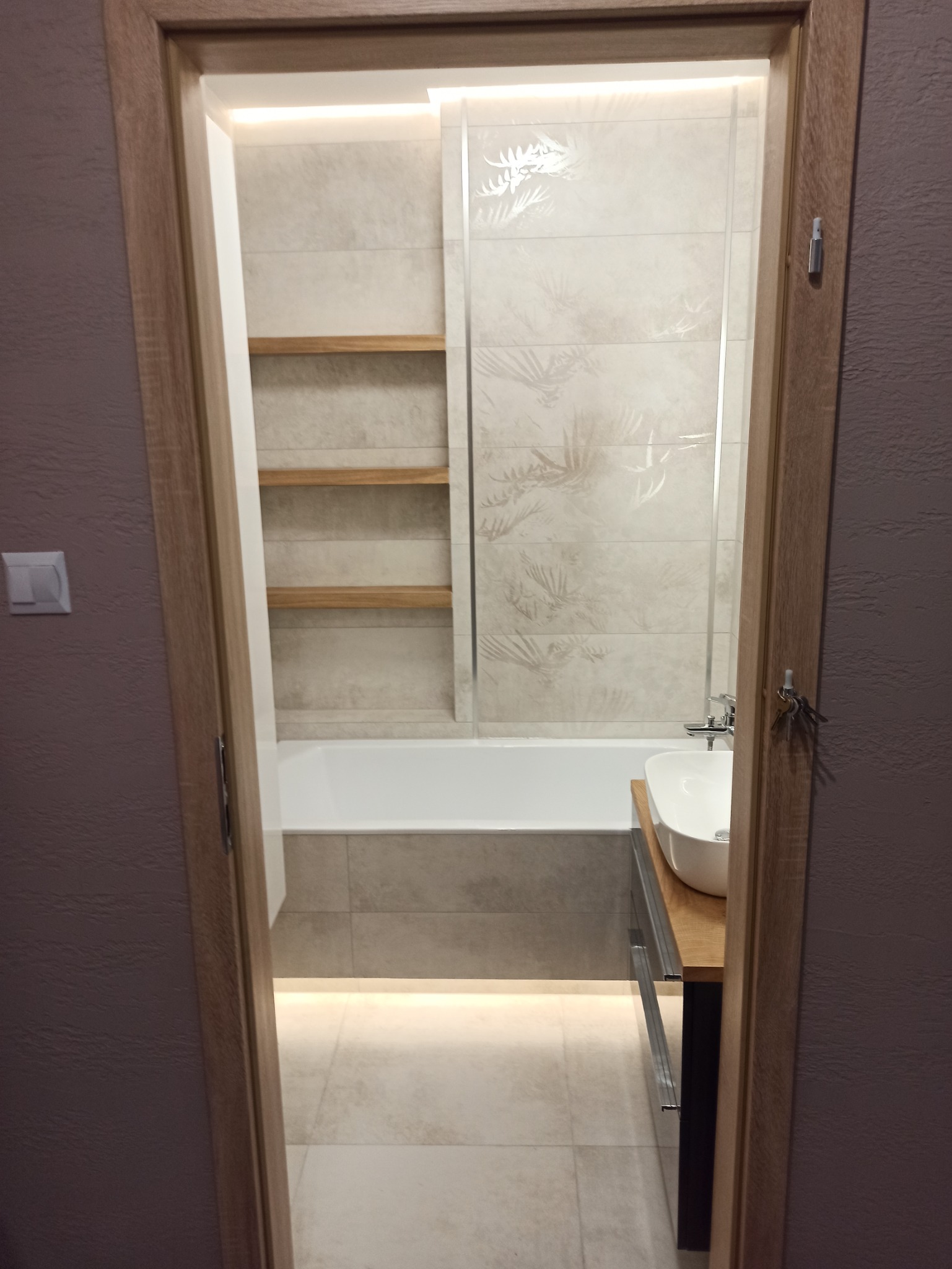 Widok na wykończoną łazienkę przez otwarte drzwi: wanna z obudową z płytek, umywalka na drewnianym blacie, płytki z motywem roślinnym na ścianie, drewniane półki w zabudowie, podświetlenie LED...